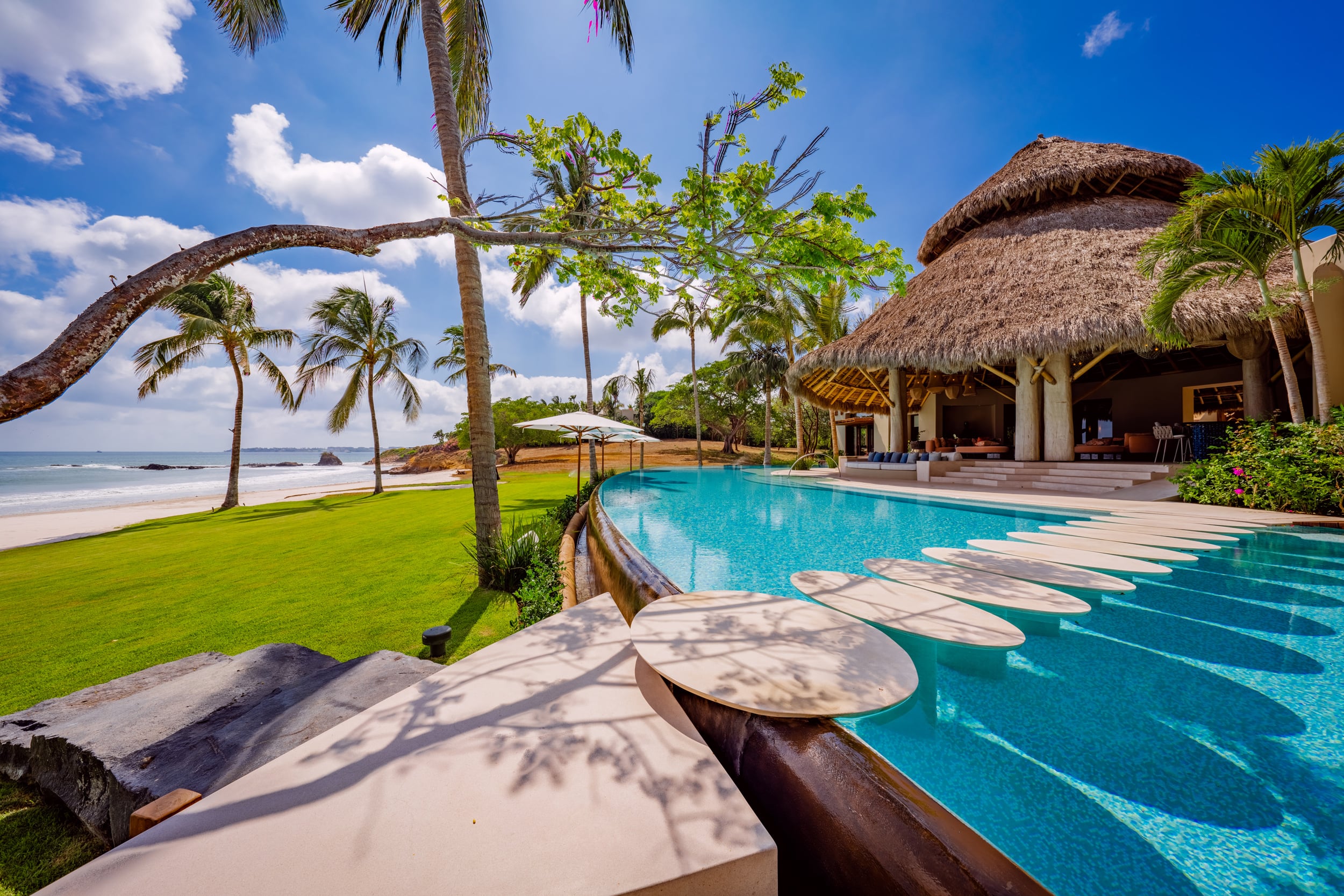 Daylight view along the pool edge where a row of oval white stepping-stone pads trails beside the water, with the palapa pavilion at the far end, beach and rocks visible to the left under a clear blue sky, Casa Akama, Ranchos Estates, Punta Mita.