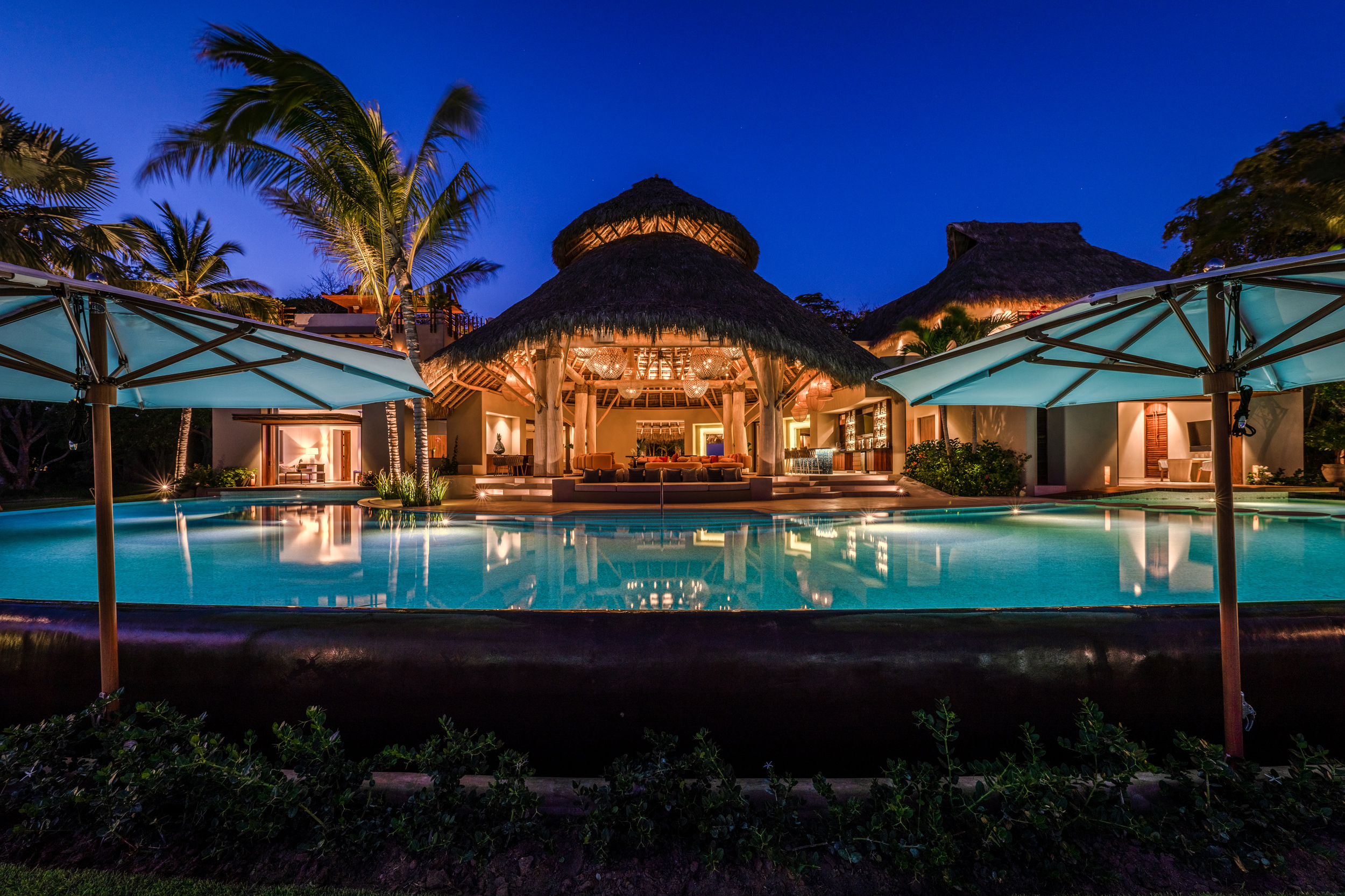Blue-hour exterior view of the estate with the pool in the foreground reflecting the illuminated palapa pavilion and lighted bedroom wing behind it, flanked by two large white umbrellas with uplights, Casa Akama, Ranchos Estates, Punta Mita.