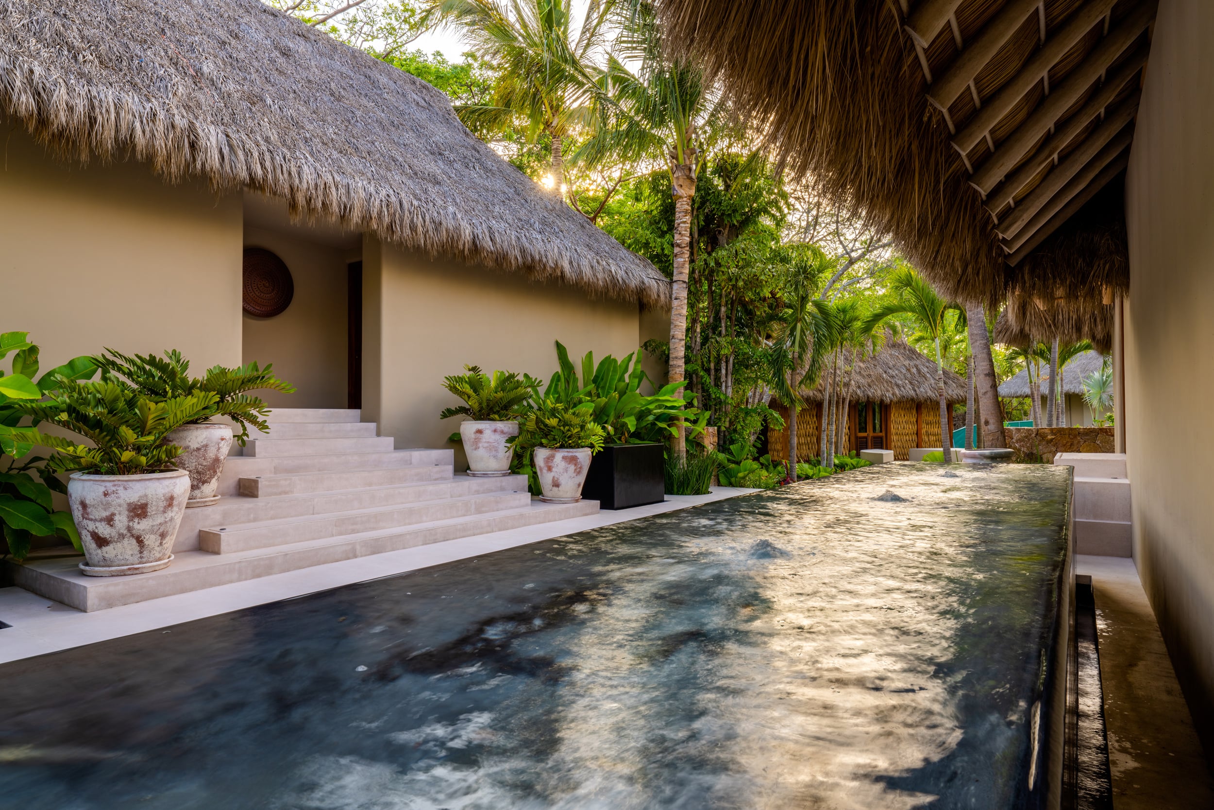 Garden pathway beside a long dark reflecting channel with still water, stone steps leading up to a palapa-roofed guest bungalow flanked by tropical plants and ceramic pots, with another thatched structure visible in the background at dusk, Casa Akama, Ranchos Estates, Punta Mita.