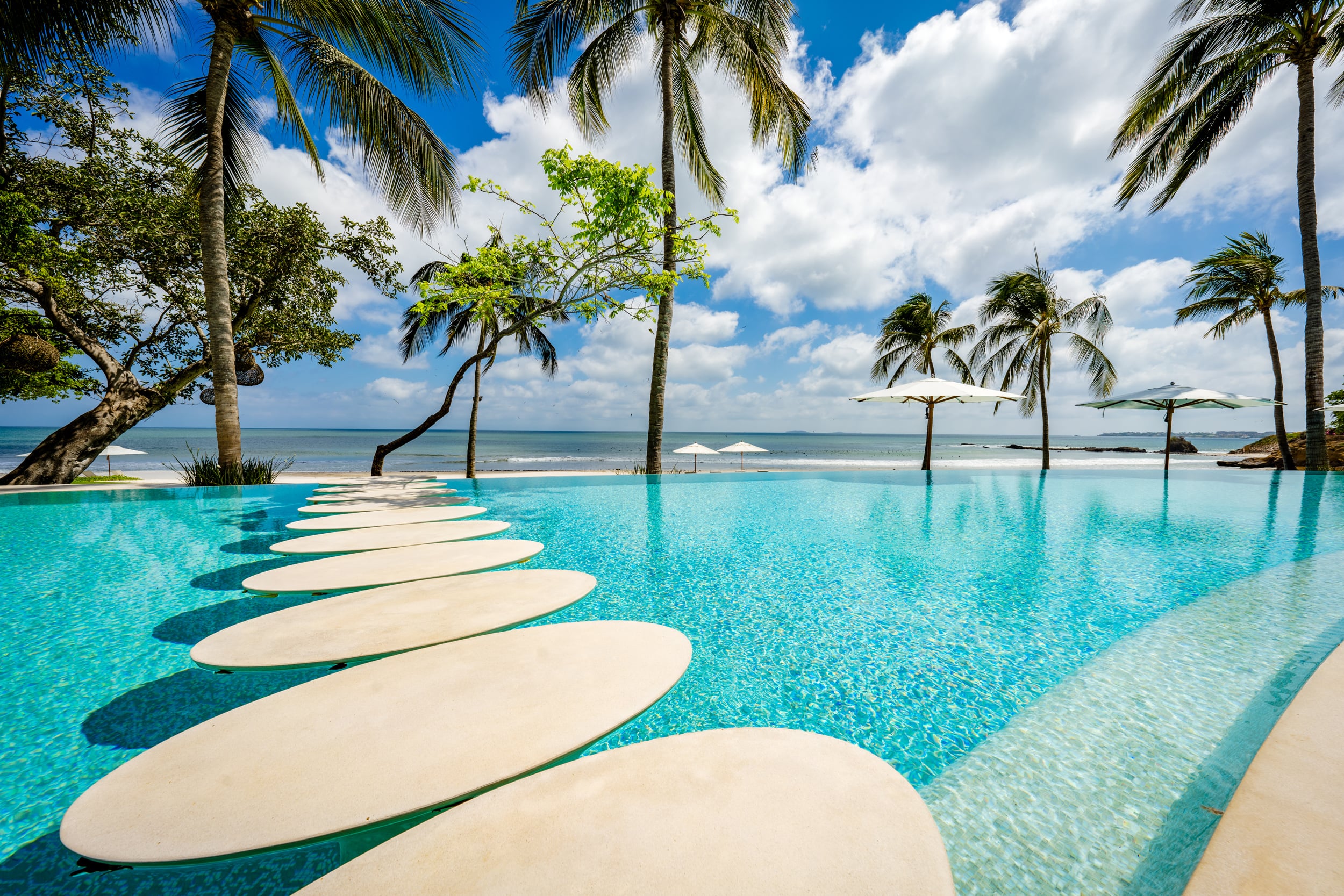Infinity pool with a row of oval white stepping pads along the inner edge, coconut palms and a broadleaf tree framing the left side, beach umbrellas on the sand, and the Pacific Ocean extending to a rocky headland under a cumulus sky, Casa Akama, Ranchos Estates, Punta Mita.