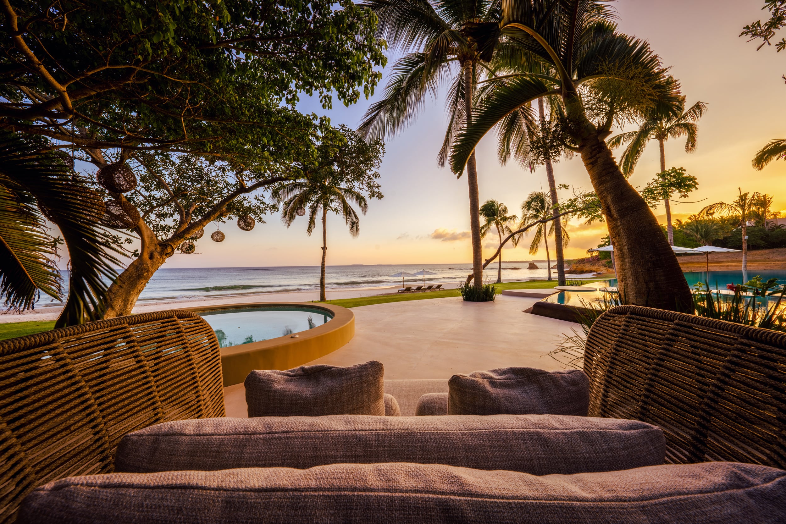 Outdoor wicker lounge chairs facing the circular plunge pool and beachfront lawn at sunset, with palm trees silhouetted against an orange sky and the main pool and palapa structure visible to the right, Casa Akama, Ranchos Estates, Punta Mita.