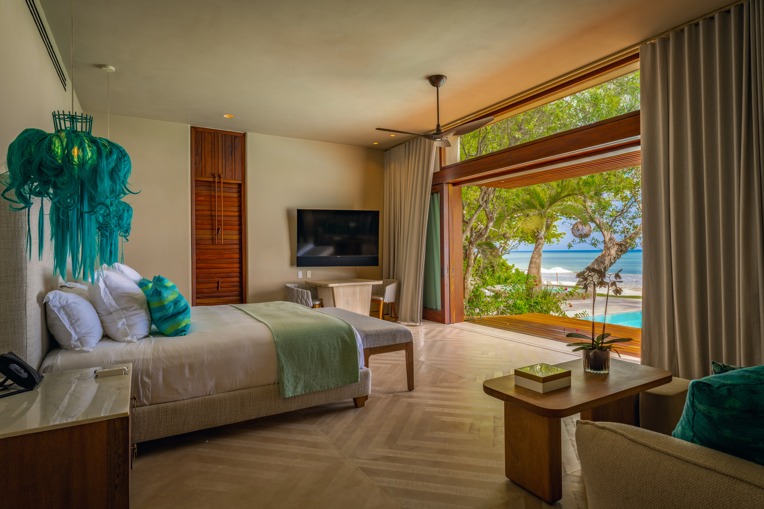 Guest bedroom with teal fiber-art chandelier, king bed with teal and green accent pillows, a floor-to-ceiling picture window framing palm trees, the pool, and the Pacific Ocean beyond, flat-screen television, and warm-wood furniture, Casa Akama, Ranchos Estates, Punta Mita.