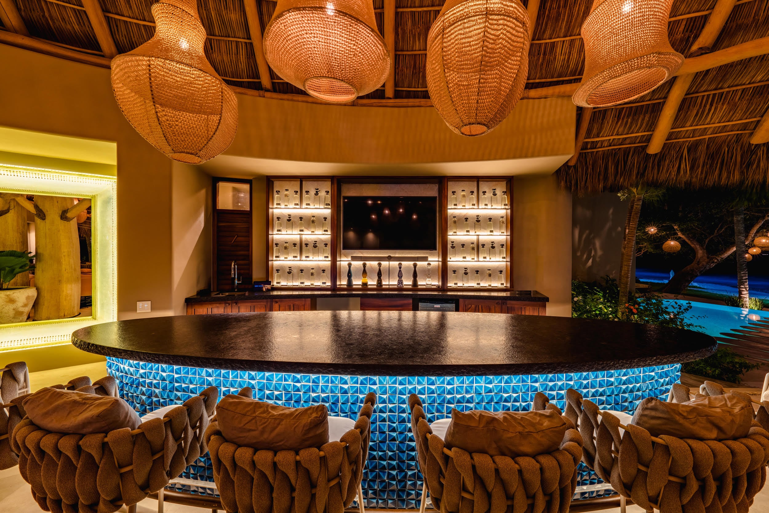 Curved palapa bar counter lit with blue tile panels, backlit bottle shelving, dark granite top, and four woven rattan barstools, with the illuminated pool visible beyond the open side, Casa Akama, Ranchos Estates, Punta Mita.