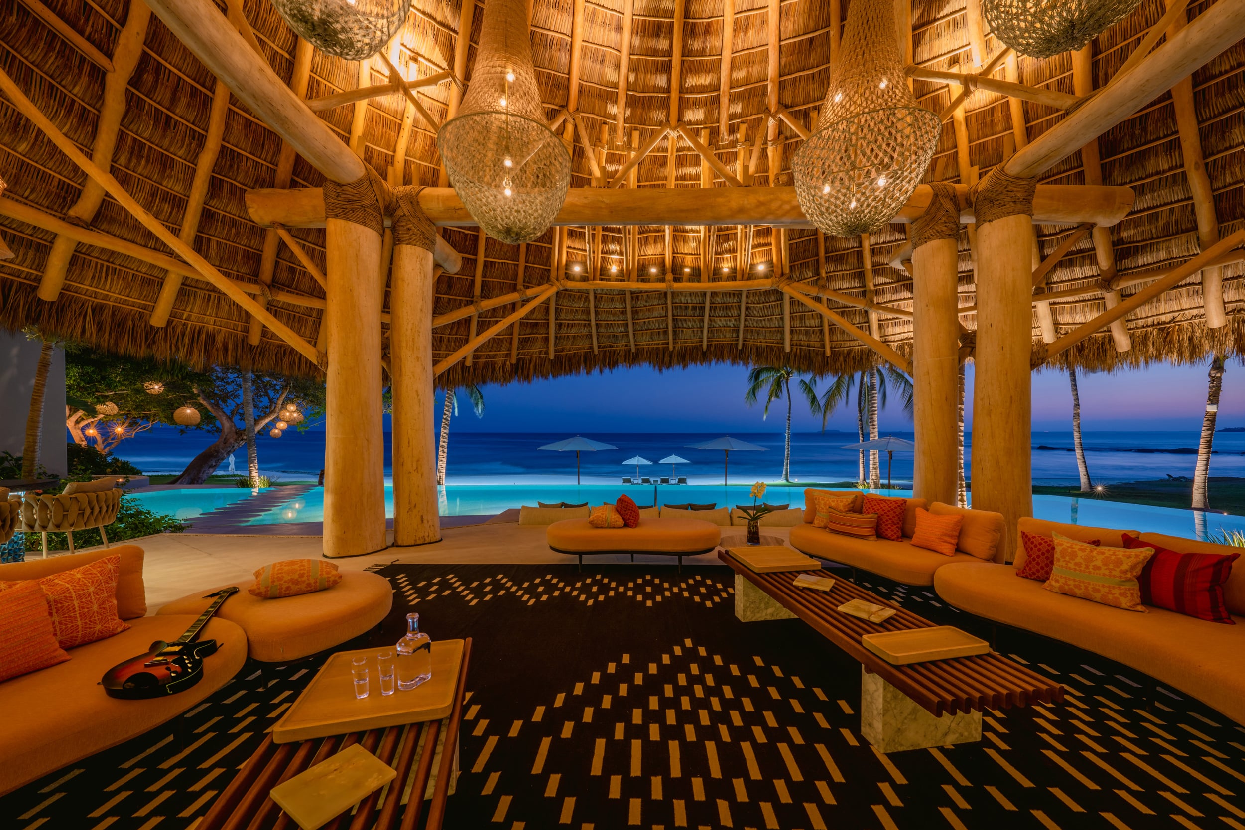 Open-sided palapa living pavilion at dusk with curved orange sofas, woven globe pendant lights, a guitar resting on the sectional, and an unobstructed view of the infinity pool and Pacific Ocean at blue hour, Casa Akama, Ranchos Estates, Punta Mita.