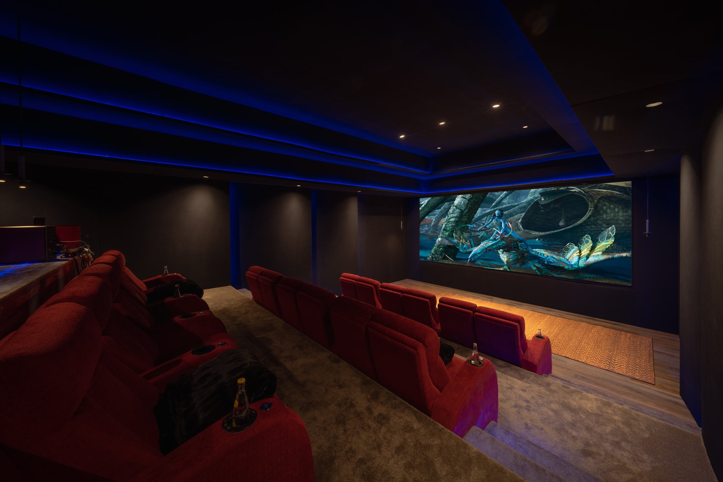 Private home cinema with three tiered rows of red velvet reclining seats, blue LED cove lighting across the ceiling, and a wide screen showing an animated film, Casa Akama, Ranchos Estates, Punta Mita.
