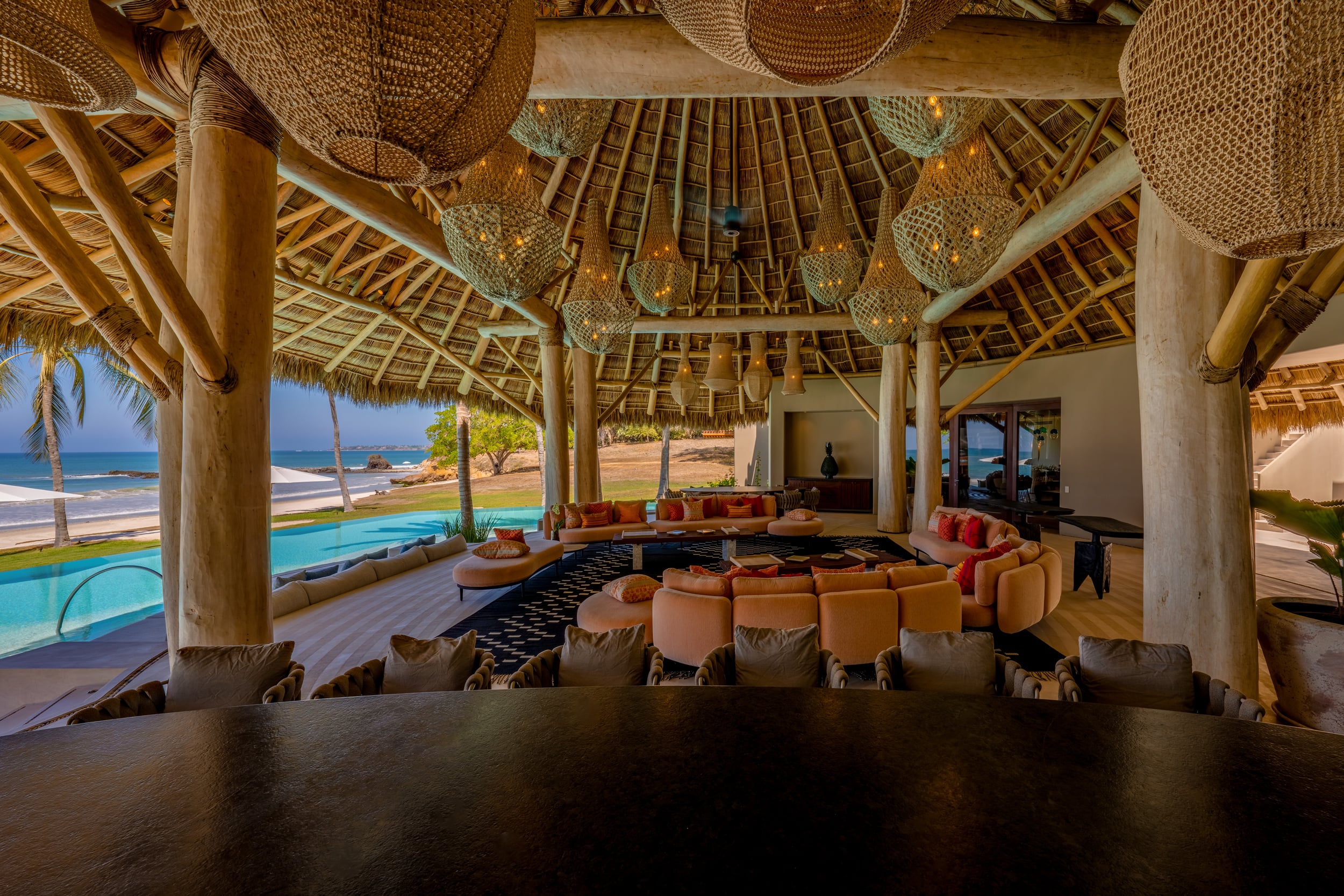 Daytime view from the dining table through the open palapa pavilion toward the circular sectional seating arrangement, the infinity pool, and the beach and ocean beyond, framed by large woven pendant lanterns overhead, Casa Akama, Ranchos Estates, Punta Mita.