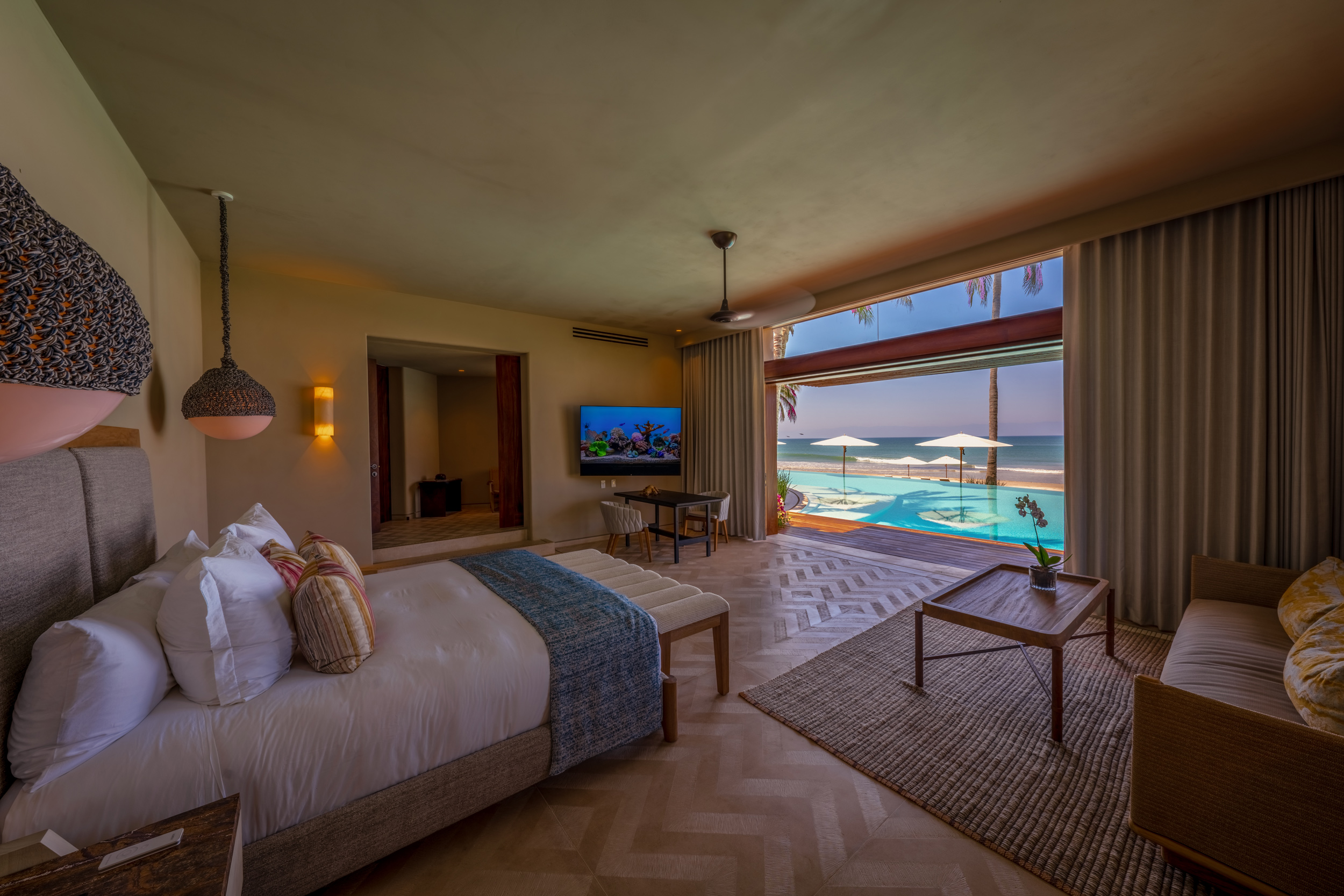 King bedroom with herringbone wood-plank floor, white upholstered headboard, woven pendant sconce, wall-mounted flat-screen TV, and open sliding glass doors framing the pool deck and Pacific Ocean with white beach umbrellas, Casa Akama, Ranchos Estates, Punta Mita.