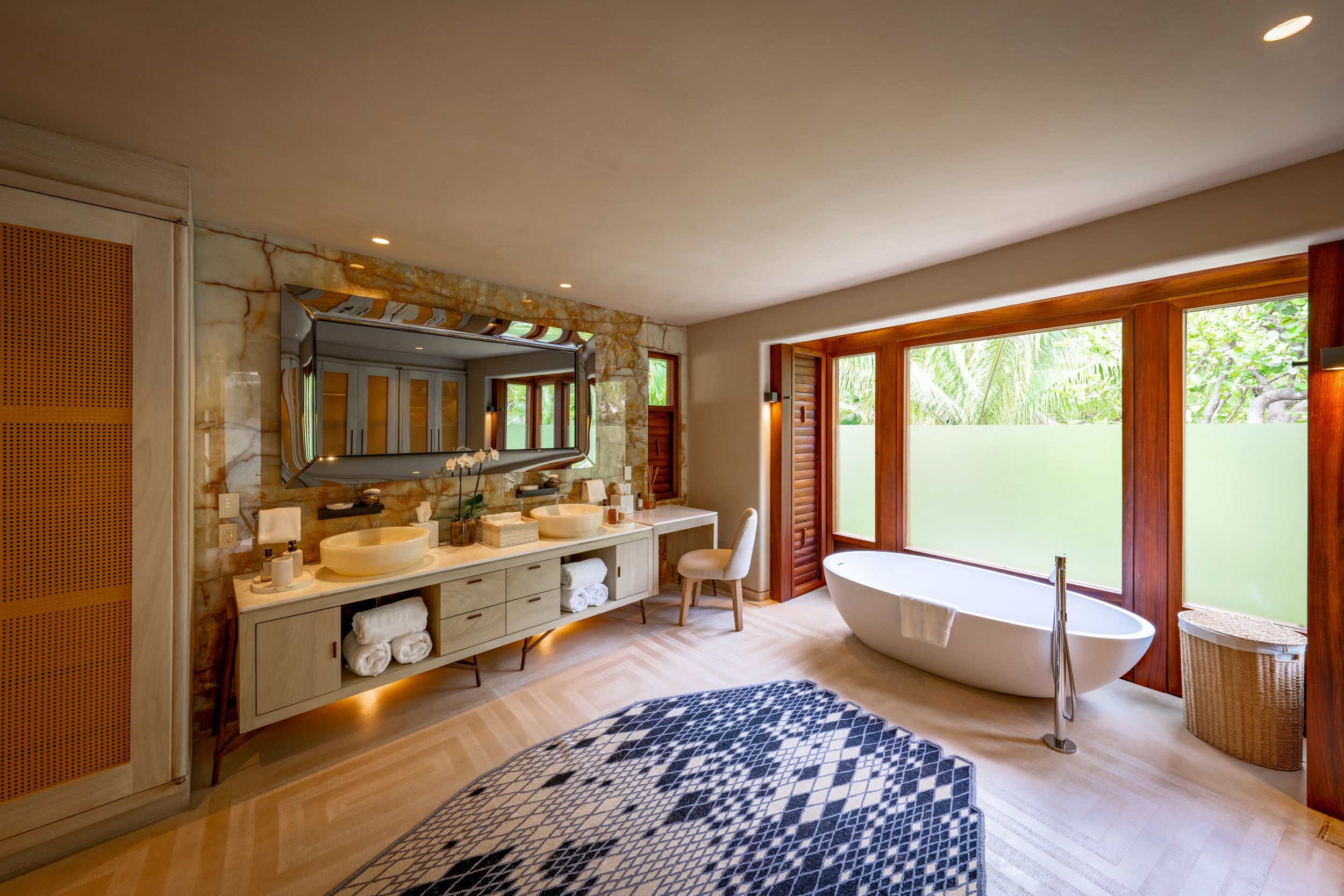 Ensuite bathroom with double vessel sinks on a stone-faced floating vanity, large framed mirror, freestanding oval soaking tub beside floor-to-ceiling teak-framed windows overlooking tropical foliage, and a diamond-pattern mosaic bath mat, Casa Akama, Ranchos Estates, Punta Mita.