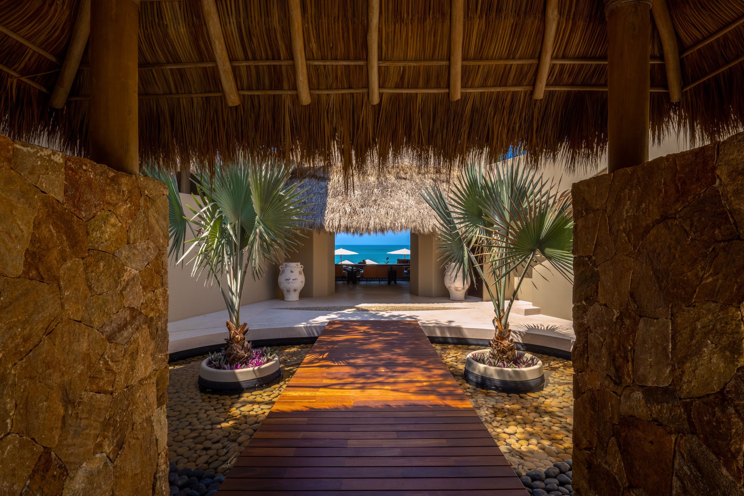Stone-walled entry corridor with a thatched palapa ceiling, a warm-toned hardwood bridge over a river-stone bed flanked by two potted fan palms, and an open view of the pool pavilion and ocean at the far end, Casa Akama, Ranchos Estates, Punta Mita.