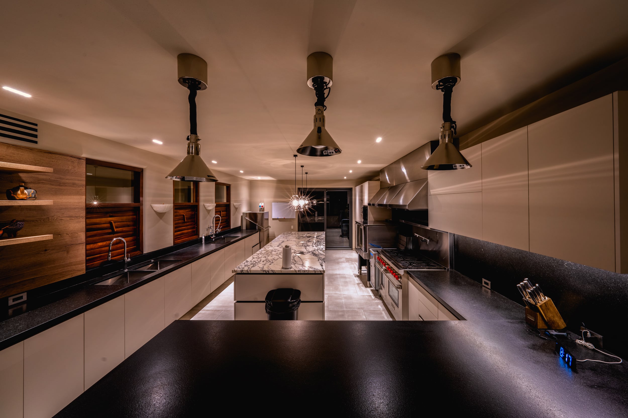 Long professional kitchen at night with twin rows of black granite counters, a central marble island, three heat-lamp pendants overhead, stainless commercial range visible mid-depth, and a knife block on the right counter, Casa Akama, Ranchos Estates, Punta Mita.