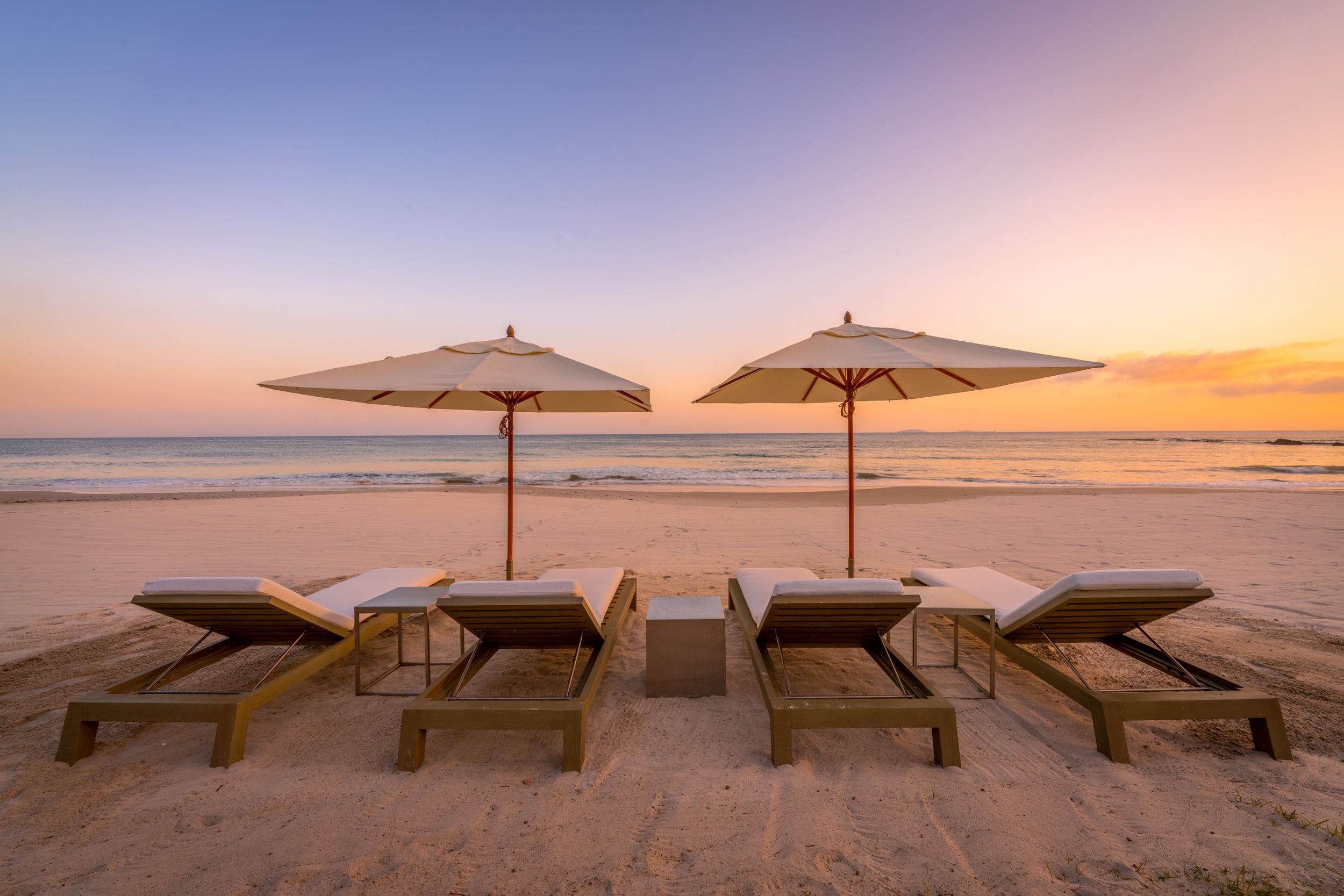Four cushioned beach loungers paired under two white canvas umbrellas on pale sand at sunset, with calm Pacific surf and a soft orange and purple sky extending to the horizon, Casa Akama, Ranchos Estates, Punta Mita.