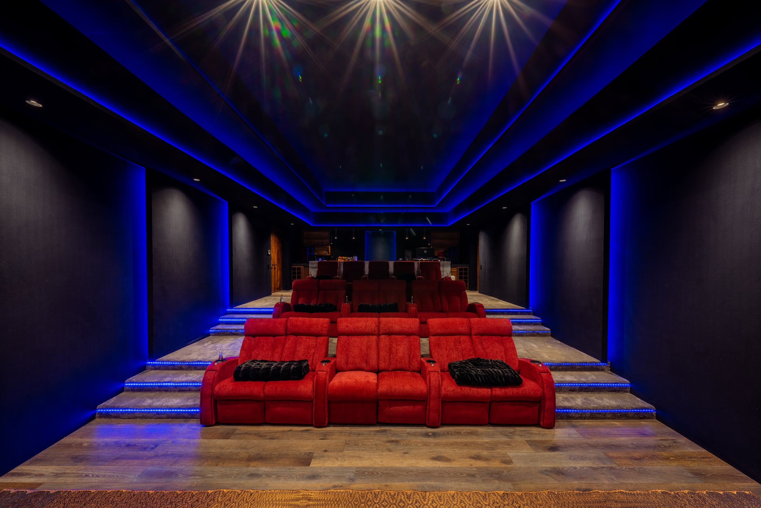 Home cinema rear view showing three stepped rows of red velvet reclining seats lit from below, blue LED perimeter lighting, star-effect ceiling lights, and a wood-plank aisle at center, Casa Akama, Ranchos Estates, Punta Mita.