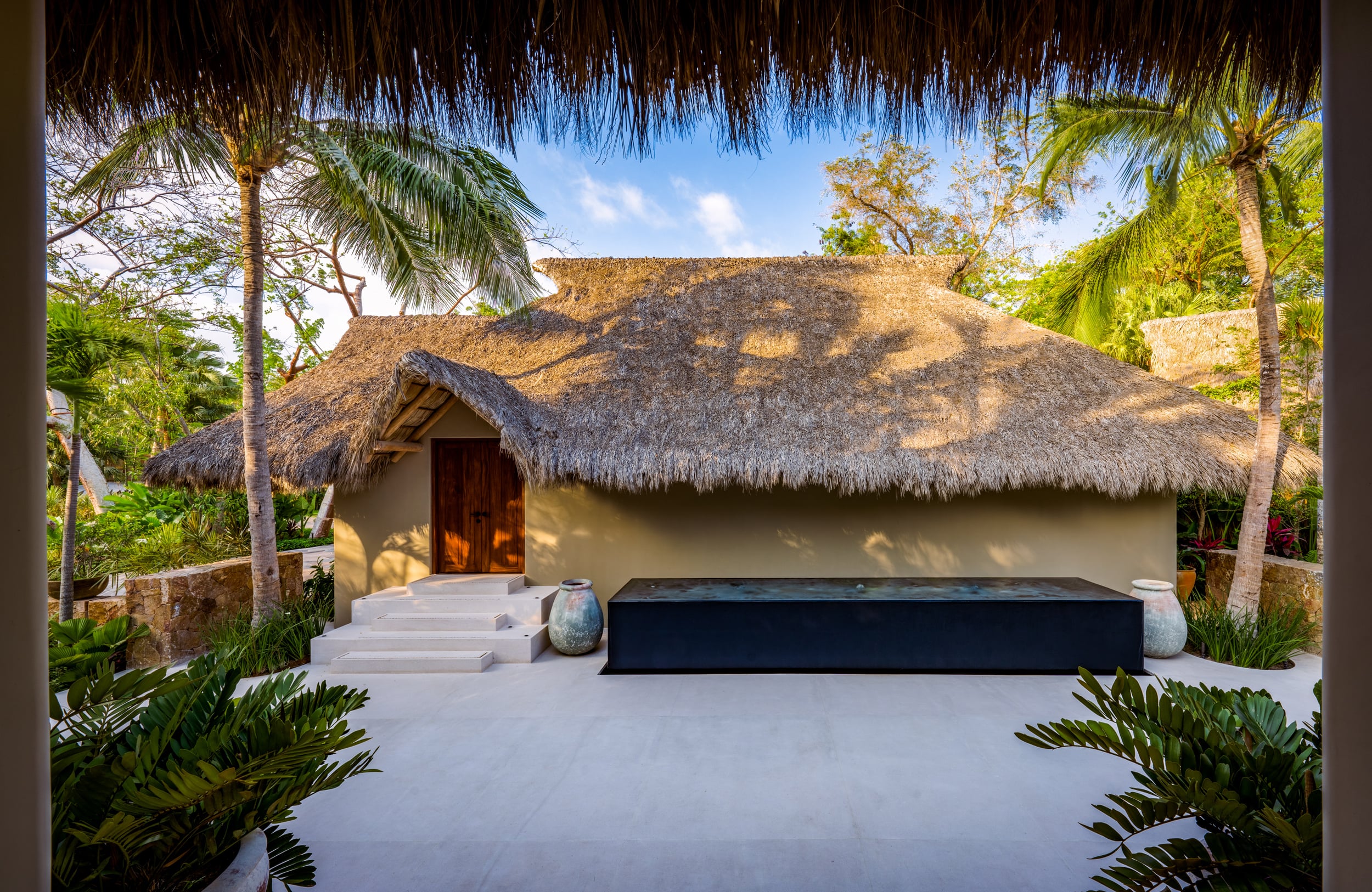 Detached thatched-roof palapa structure with a solid wooden entry door framed by steps, flanked by large ceramic urns and tropical planting, on a smooth concrete plaza, Casa Akama, Ranchos Estates, Punta Mita.