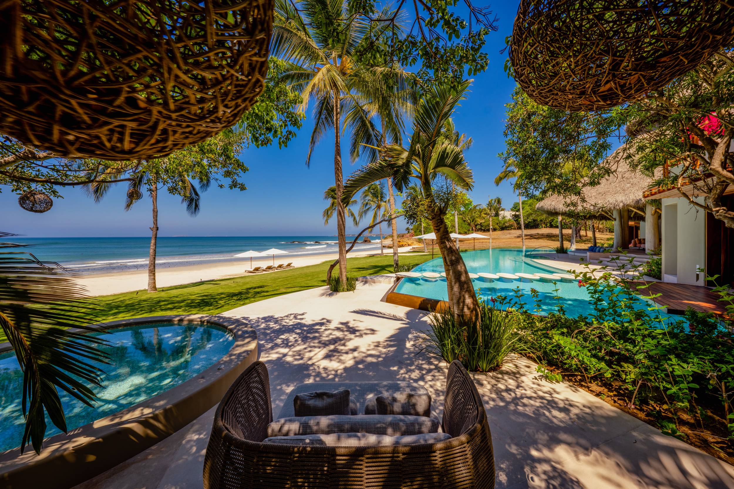 Shaded lounge area with two large woven-twig pod chairs and a sofa cushion looking out through palm and tropical canopy toward the infinity pool, lawn, beach umbrellas, and Pacific Ocean under a clear blue sky, Casa Akama, Ranchos Estates, Punta Mita.