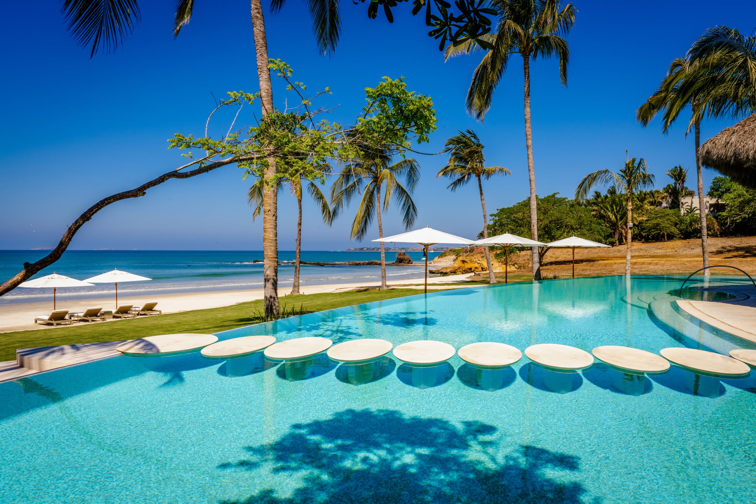 Beachfront infinity pool with a row of circular in-pool platforms along the near edge, tall palm trees behind, beach umbrellas on the lawn, and open Pacific Ocean visible beyond the pool's vanishing edge, Casa Akama, Ranchos Estates, Punta Mita.