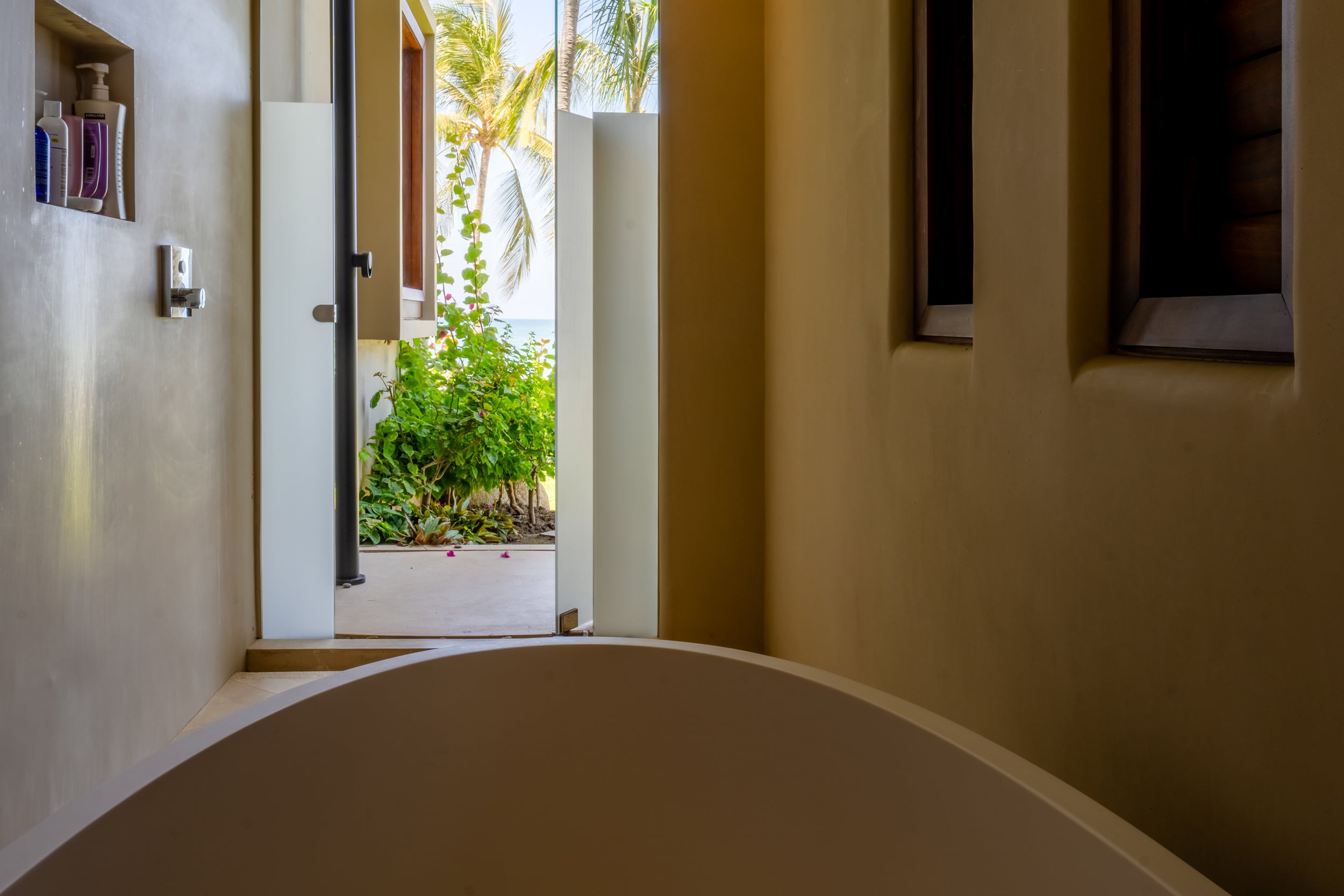Close-up ensuite bathroom view of a freestanding oval tub with an open door beyond revealing a bougainvillea-draped outdoor corridor and palm tree with ocean glimpsed at top, toiletry bottles visible in a niche, Casa Akama, Ranchos Estates, Punta Mita.