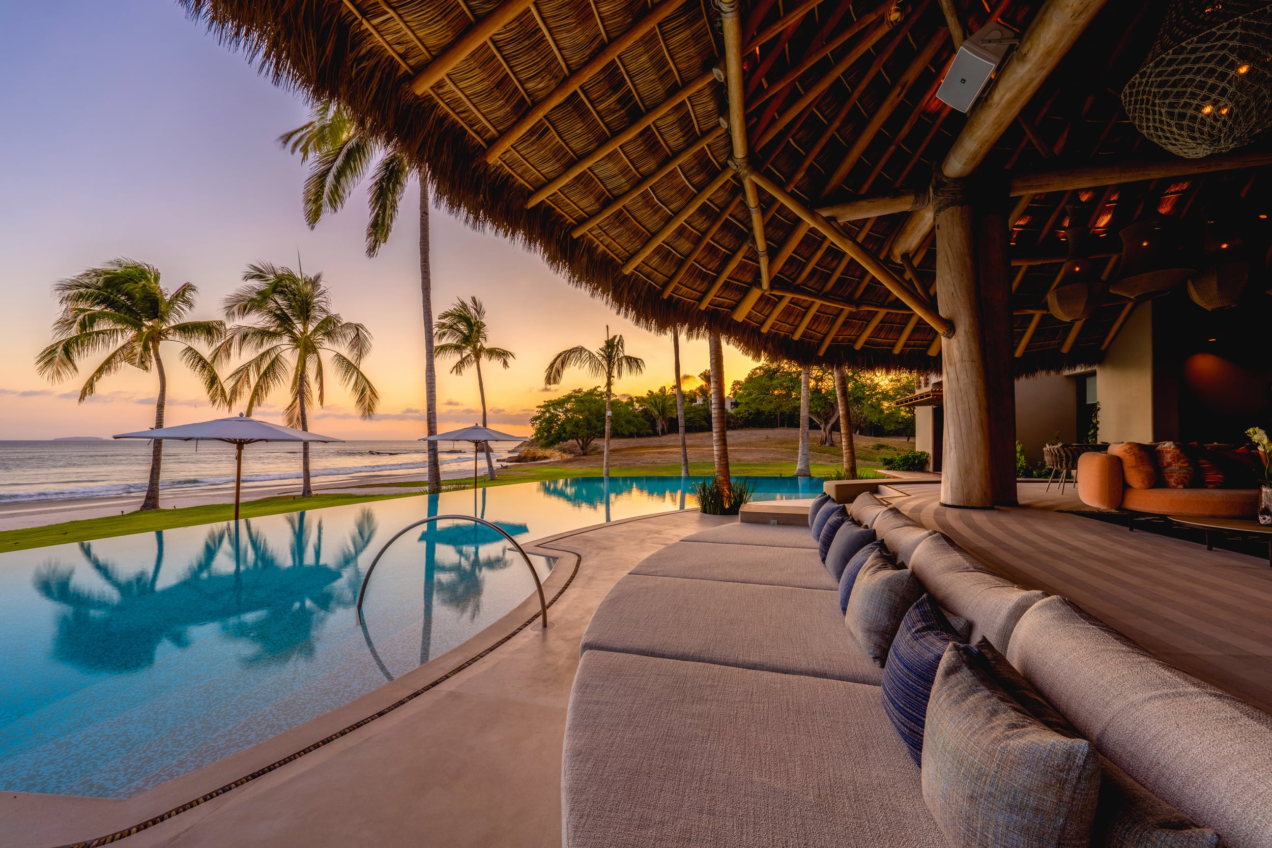 Infinity pool and palapa pavilion at sunset, a long cream sectional sofa running along the palapa edge, the pool reflecting warm sky tones, coconut palms silhouetted against orange and pink dusk light over the ocean, Casa Akama, Ranchos Estates, Punta Mita.