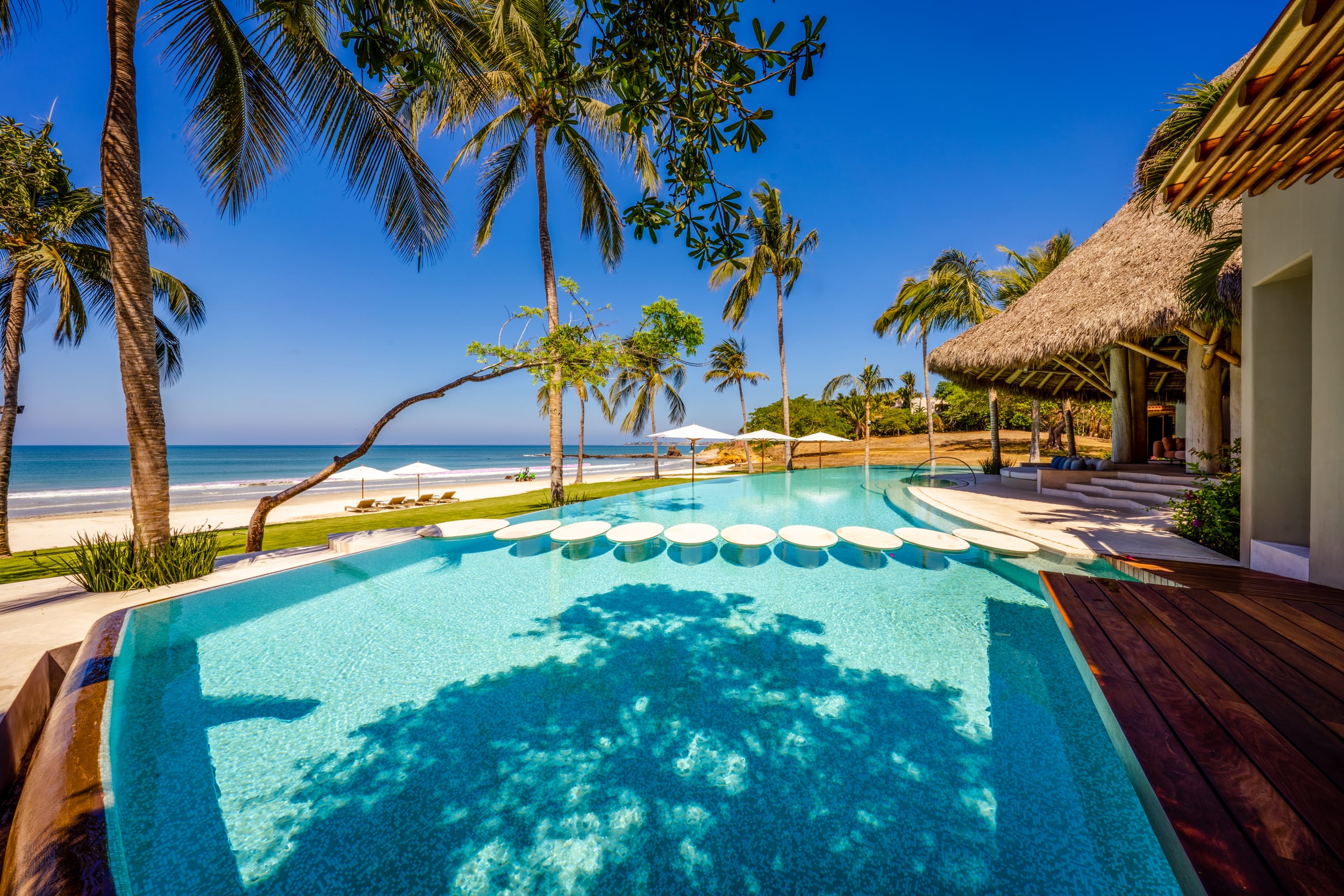 Daytime shot of the beachfront infinity pool from the teak deck, circular in-pool stepping platforms visible along the near edge, beach umbrellas on the lawn, palm trees framing the view, and open Pacific water beyond, Casa Akama, Ranchos Estates, Punta Mita.