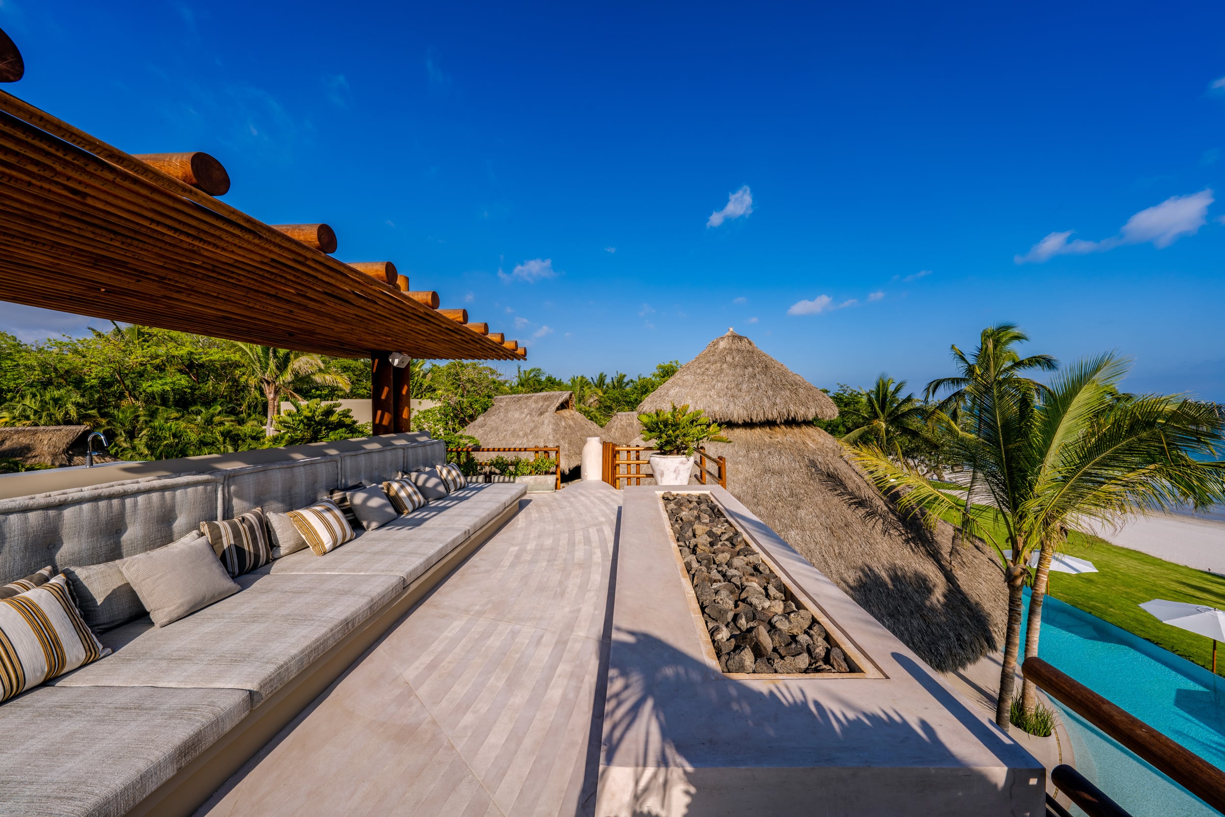 Upper-level rooftop terrace with a long built-in grey sectional sofa under a slatted wood pergola overhang, a narrow volcanic-rock fire channel at center, thatched pavilion rooftops at mid-ground, and ocean visible in the distance, Casa Akama, Ranchos Estates, Punta Mita.