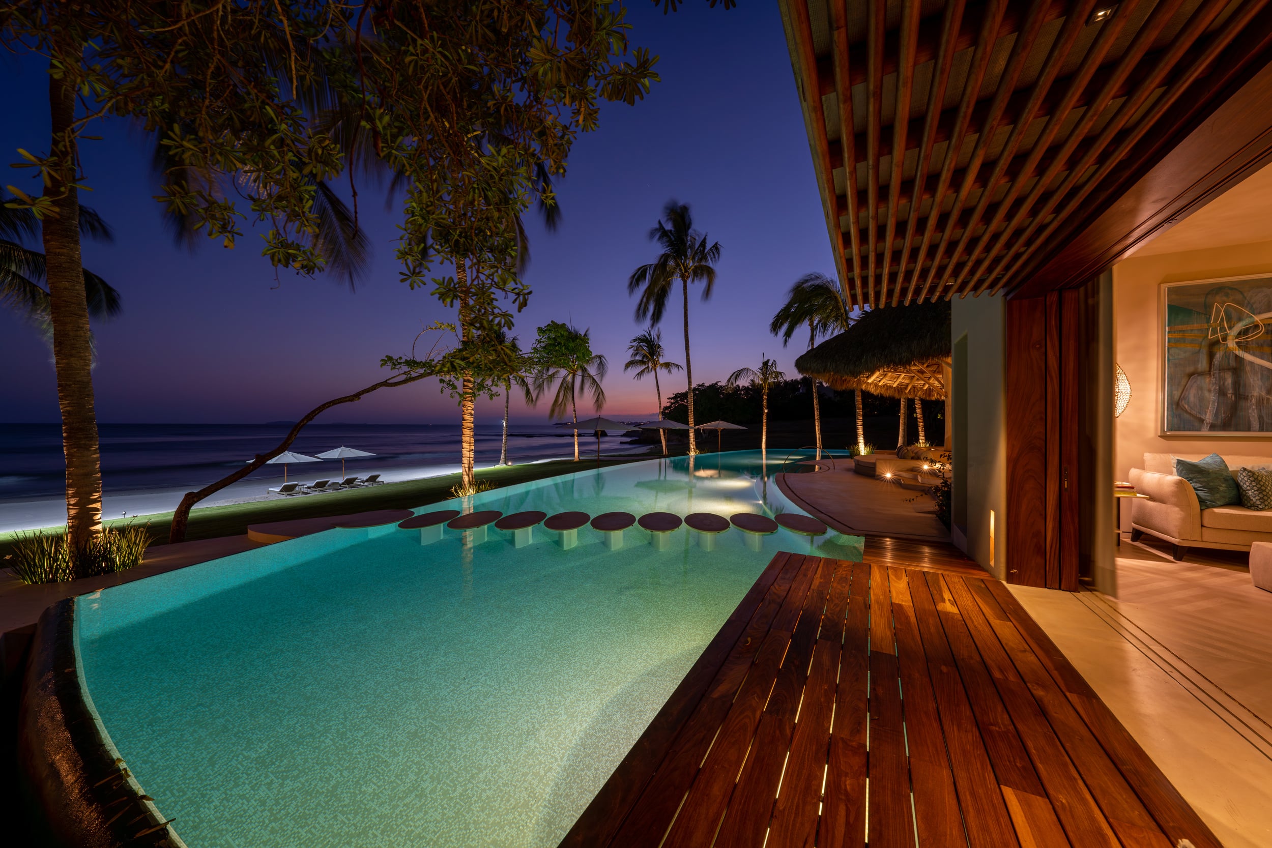 Beachfront infinity pool illuminated at night from the teak deck, circular stepping pads glowing beneath the water surface, interior room art-lit through open sliding doors at right, palms and beach umbrella silhouetted against deep blue dusk, Casa Akama, Ranchos Estates, Punta Mita.