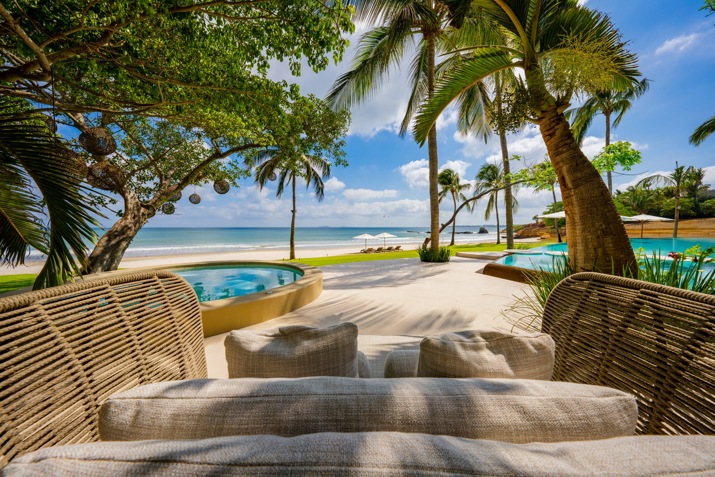 Garden lounge with two large woven rattan chairs and a neutral linen cushion in the foreground, facing an open pool terrace with palm canopy, a circular plunge pool in the mid-ground, beach umbrellas, and Pacific Ocean under a clear sky, Casa Akama, Ranchos Estates, Punta Mita.