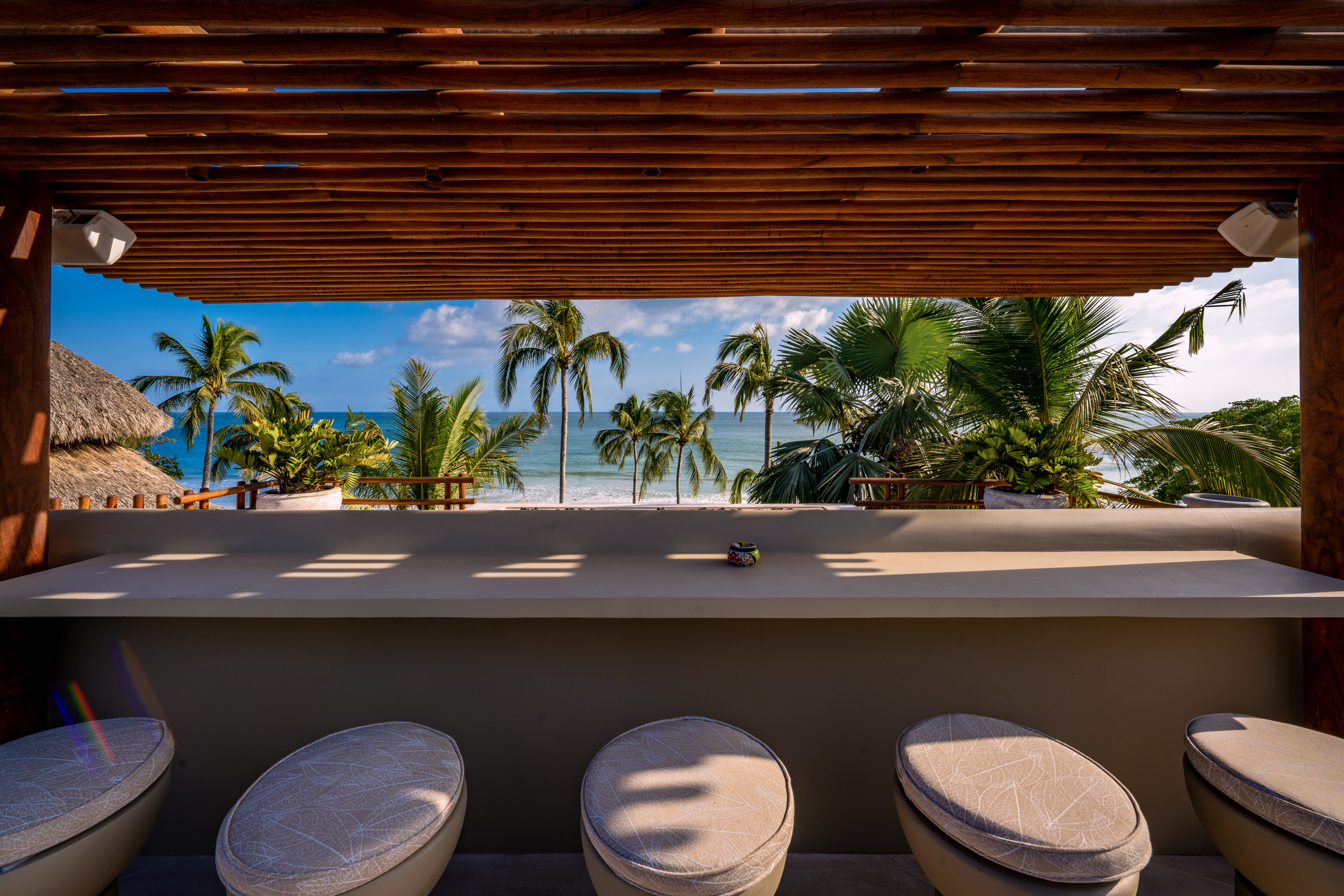 Rooftop bar area viewed from behind three round cushioned barstools, a wide concrete counter stretching toward an opening framed by slatted wood beams, with palms, the pavilion rooftop, and the Pacific Ocean filling the view, Casa Akama, Ranchos Estates, Punta Mita.