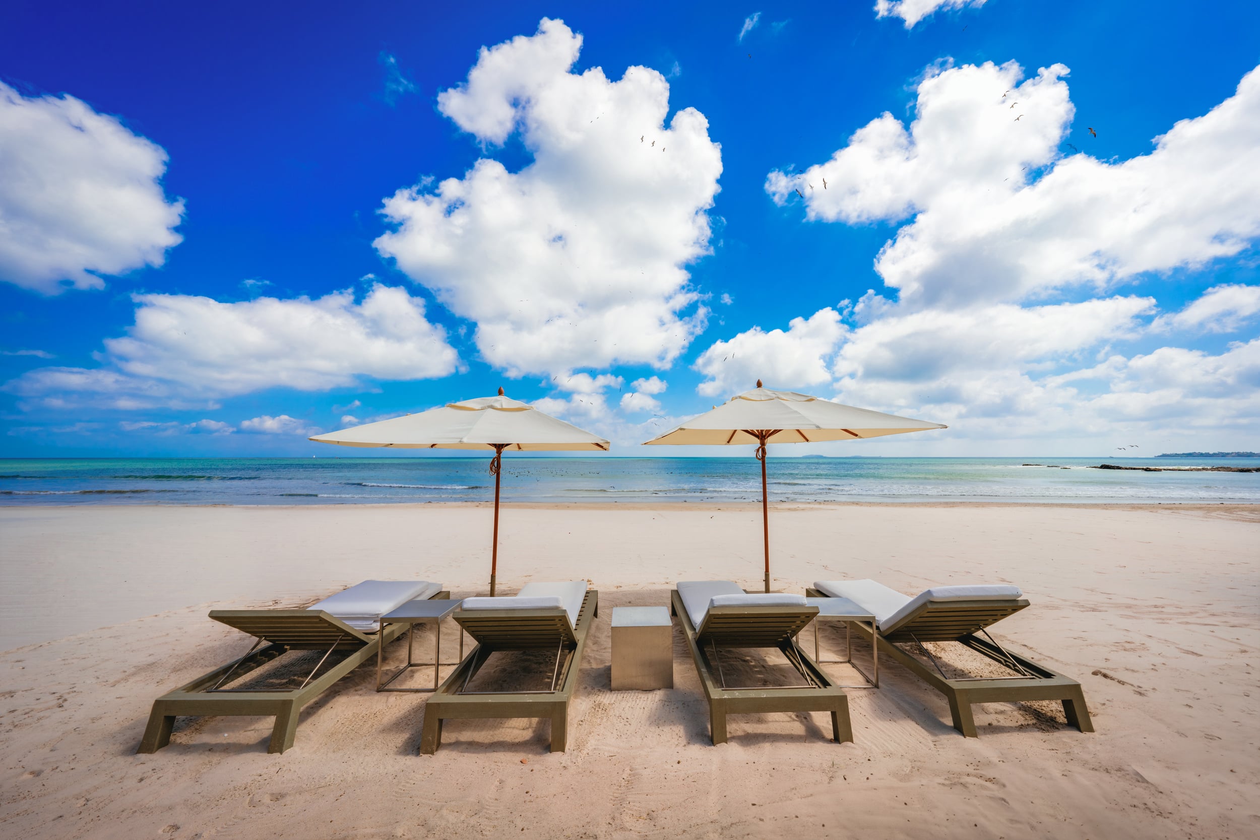 Four padded beach loungers with two white canvas umbrellas arranged on white sand under a bright blue sky with cumulus clouds, the calm turquoise Pacific Ocean visible to the horizon, Casa Akama, Ranchos Estates, Punta Mita.