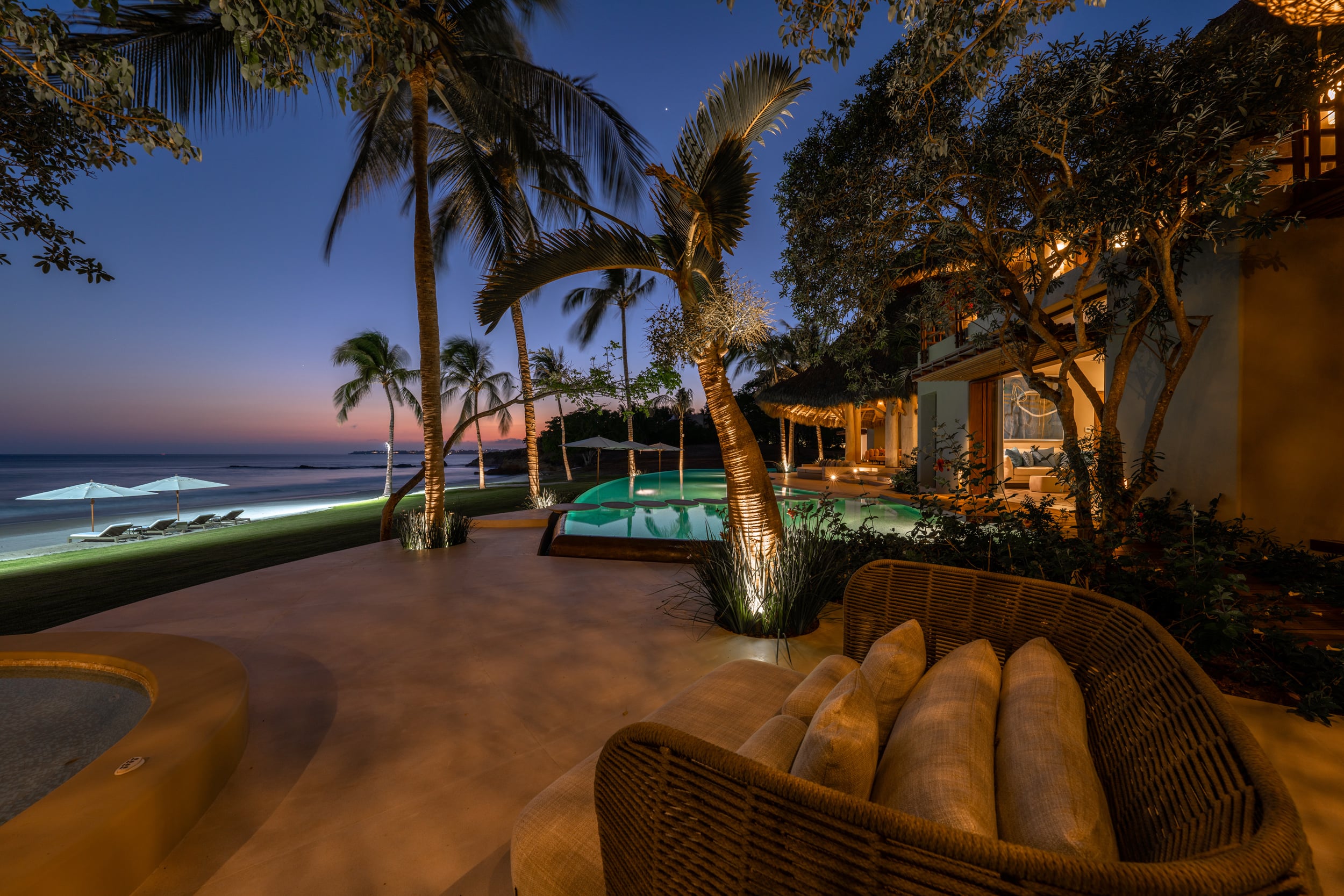 After-dark terrace view with a wicker lounge chair in the foreground, the lit infinity pool and circular plunge pool in the mid-ground, palm trees uplighted, and the villa pavilion glowing warmly against a deep blue post-sunset sky, Casa Akama, Ranchos Estates, Punta Mita.