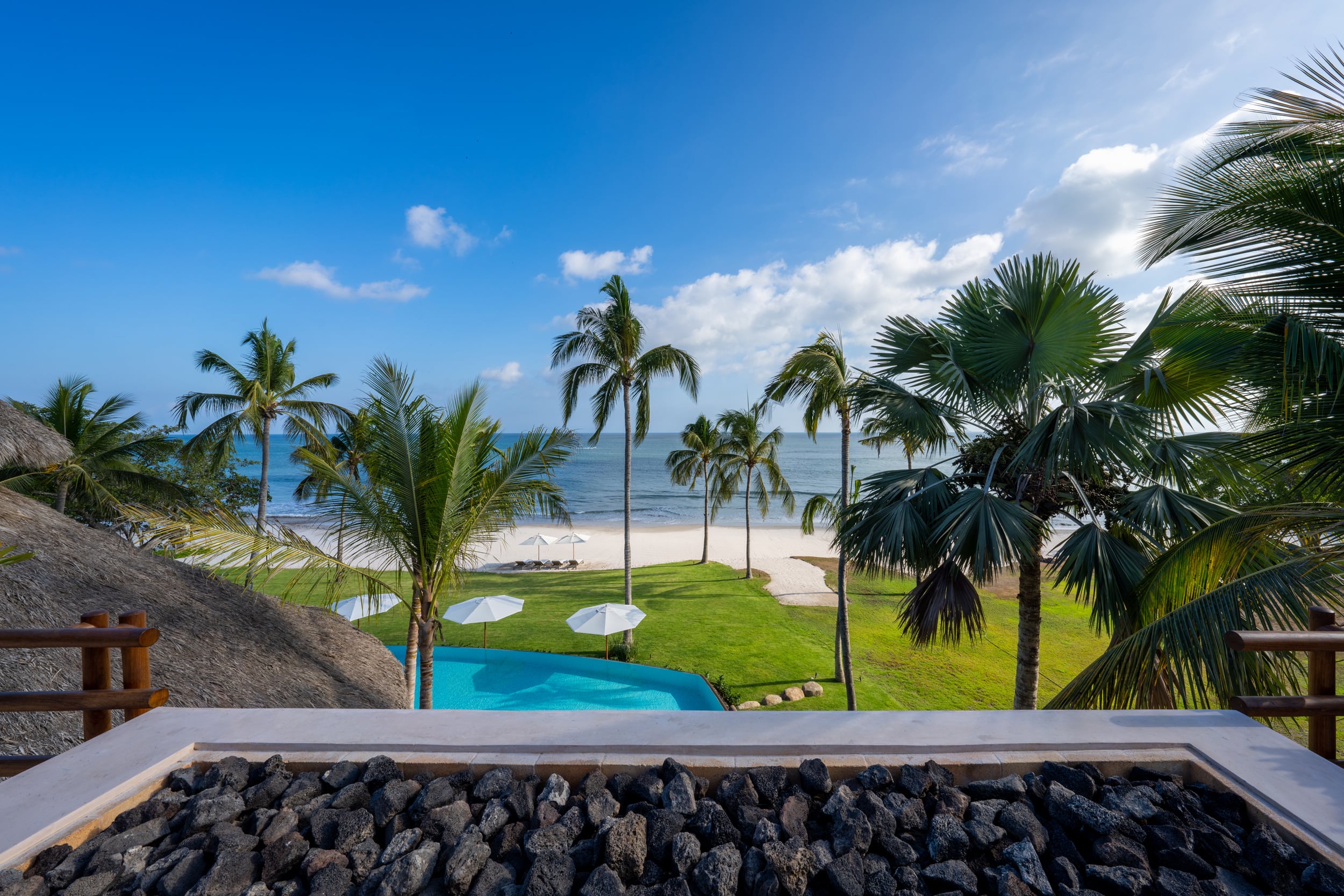 Elevated view from a rooftop or upper balcony overlooking the lawn, a small rectangular pool with beach umbrellas, coconut palms and fan palms, sandy beach, and the open Pacific Ocean under a clear blue sky, Casa Akama, Ranchos Estates, Punta Mita.