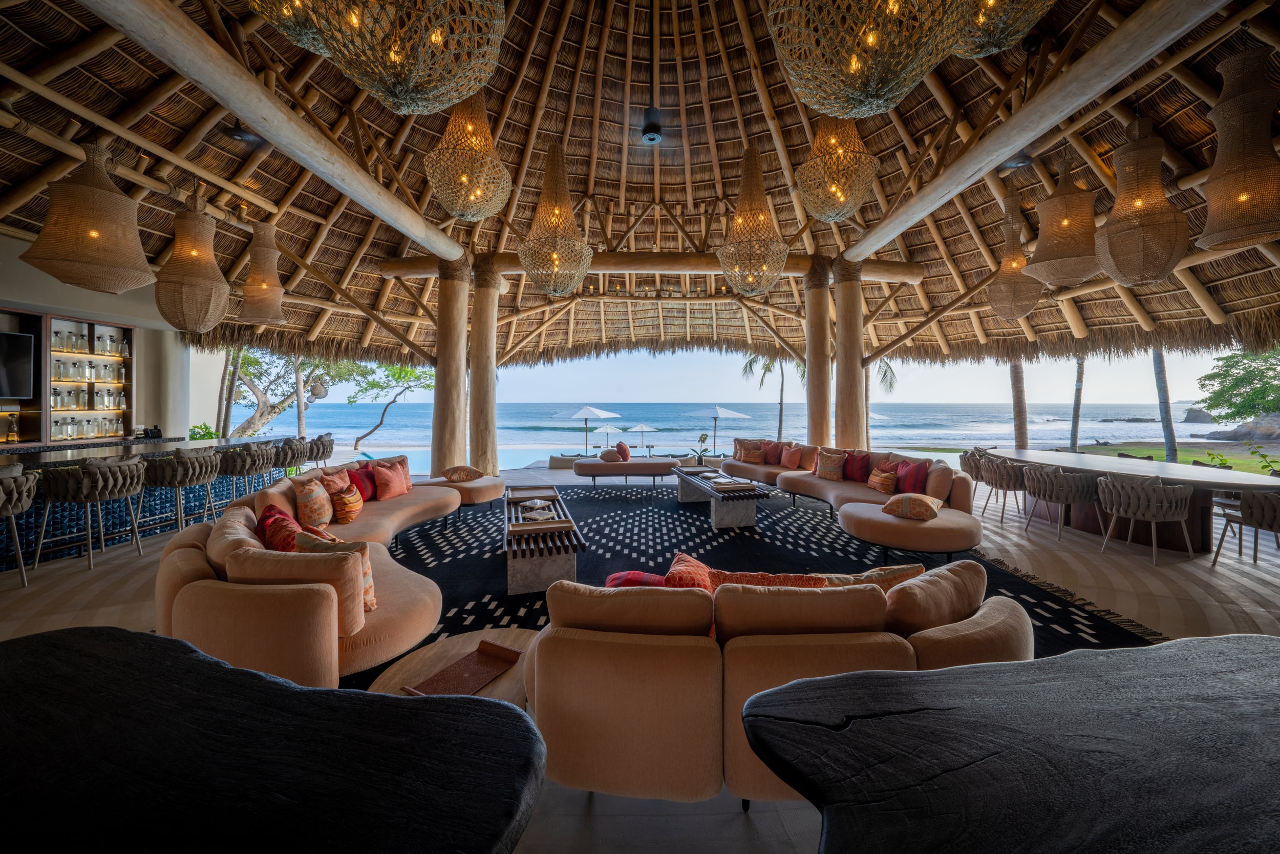 Wide palapa living pavilion interior with cream curved sectional sofas, woven rattan pendant lanterns, a thatched palapa ceiling spanning a large circular plan, bar seating with blue tile to the left, and an open beachfront view through the pavilion columns, Casa Akama, Ranchos Estates, Punta Mita.