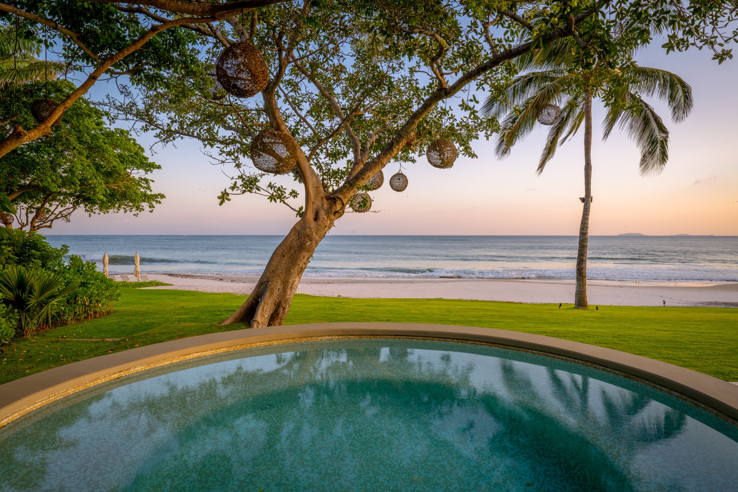 Circular plunge pool at dusk with a large shade tree hung with woven rattan lanterns, coconut palms framing the left side, a green lawn meeting pale sand, and soft orange and pink tones at the horizon over the Pacific Ocean, Casa Akama, Ranchos Estates, Punta Mita.