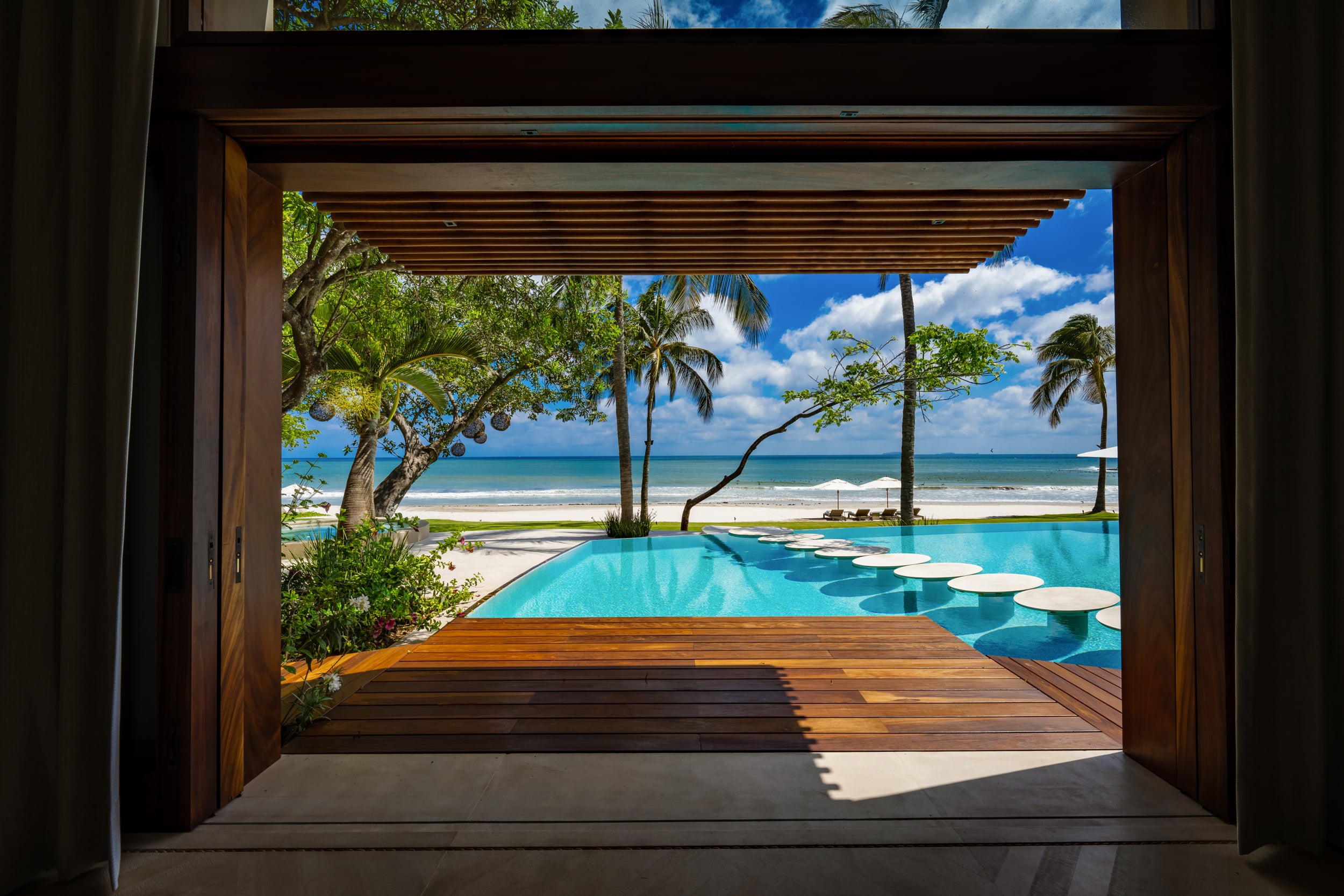 Interior view looking out through wide open teak-framed sliding doors across a hardwood deck to the infinity pool with circular stepping pads, lawn, beach umbrellas, palm canopy, and the Pacific Ocean under a cloud-dotted blue sky, Casa Akama, Ranchos Estates, Punta Mita.