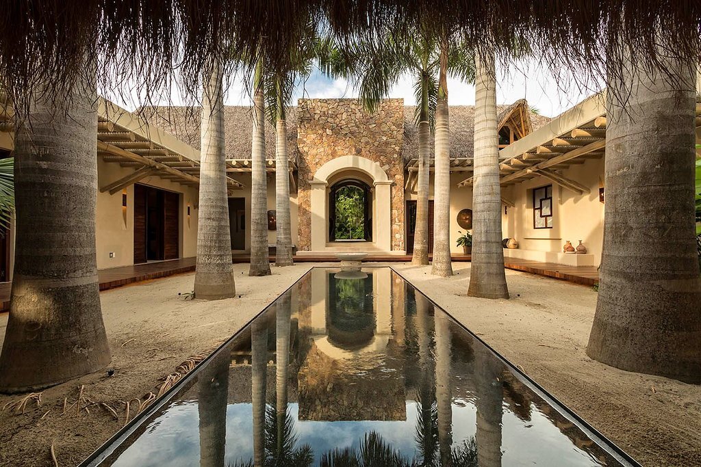 Symmetrical courtyard reflecting pool flanked by tall royal palms and a thatched palapa roof, sand underfoot and an arched stone entryway ahead, Casa Escondida, Ranchos Estates, Punta Mita.