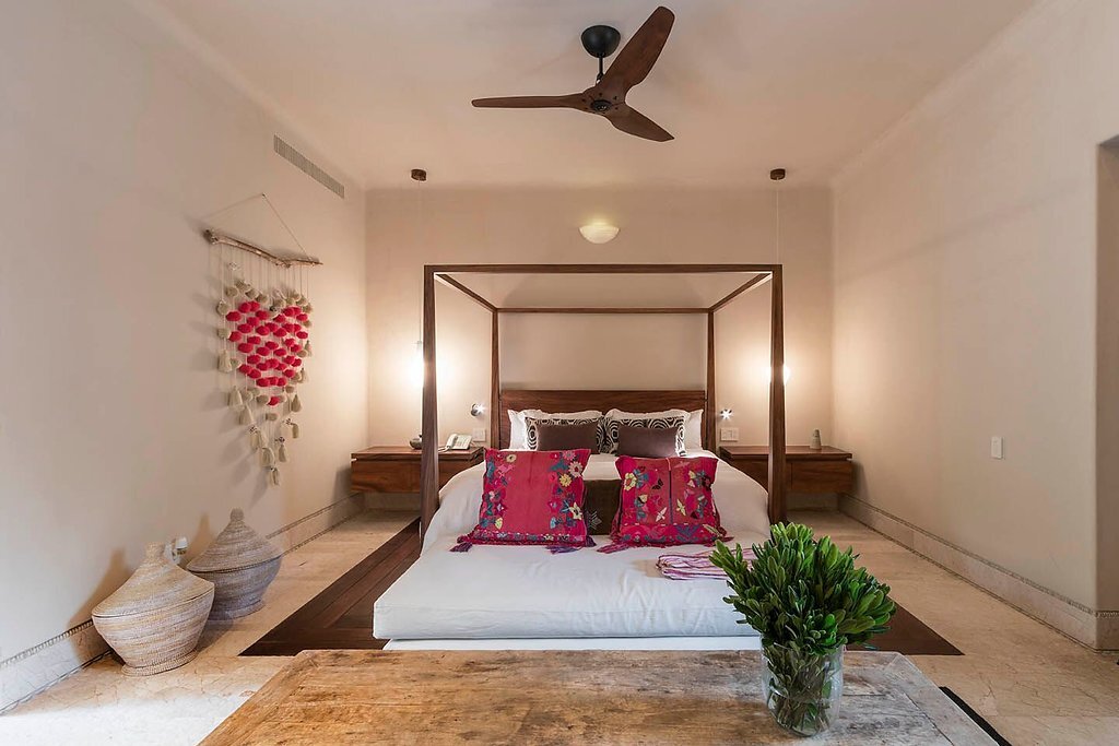 Four-poster king bed with embroidered pink pillows and white linens, shell wall hanging, travertine floor, and ceiling fan in a warm candlelit suite, Casa Escondida, Ranchos Estates, Punta Mita.
