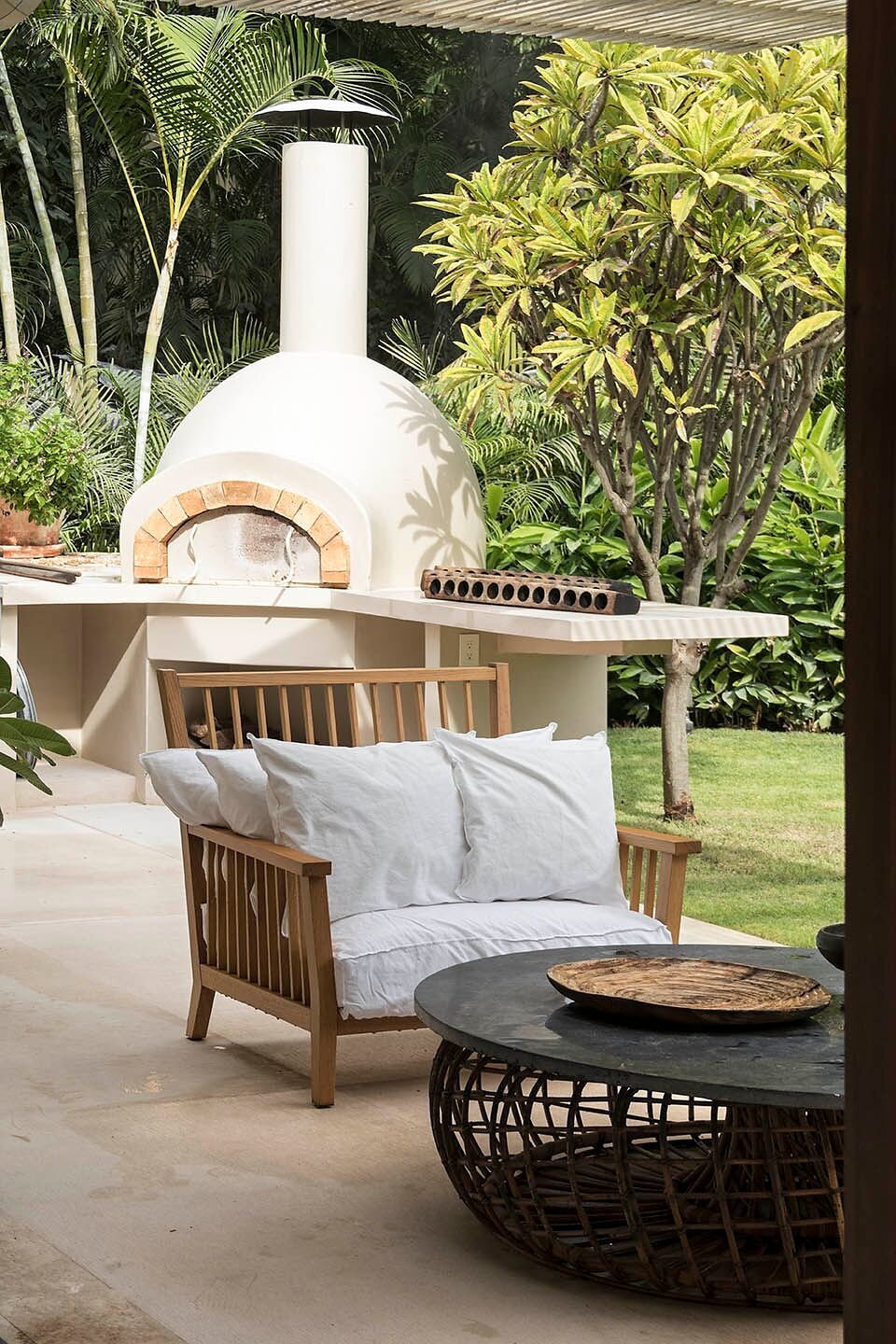 Outdoor terrace with a white wood-fired pizza oven framed by brick, a teak sofa with white cushions, and a round wicker table set against a lush tropical garden, Casa Escondida, Ranchos Estates, Punta Mita.