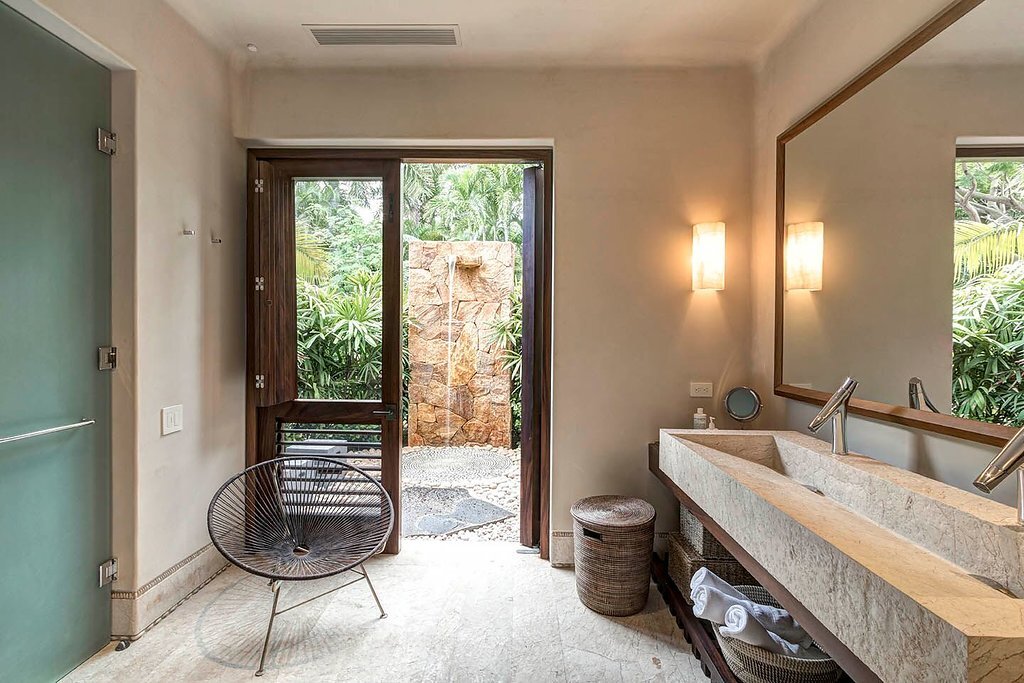 Ensuite bath with a long travertine trough sink, warm wall sconces, an Acapulco chair, and a glass door opening to a private outdoor stone shower in the tropical garden, Casa Escondida, Ranchos Estates, Punta Mita.