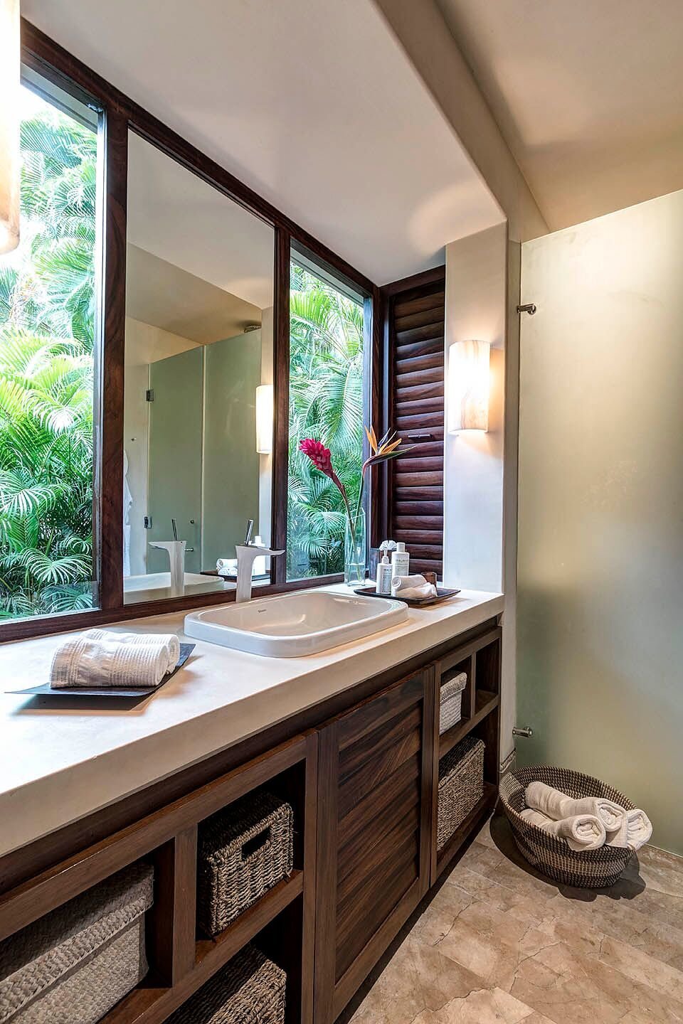 Dark wood vanity with a white stone countertop, large garden-view window, woven basket storage below, a single bird-of-paradise bloom, and wall sconce lighting, Casa Escondida, Ranchos Estates, Punta Mita.