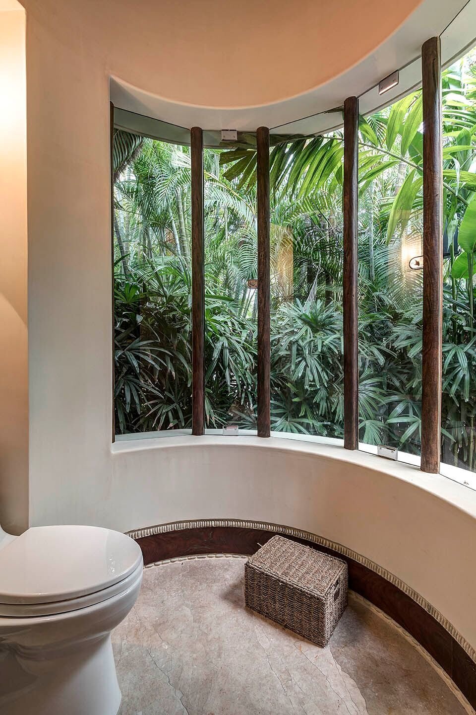 Curved bathroom with a floor-to-ceiling arc of windows looking into a dense palm garden, travertine floor, and a small woven stool beside the toilet, Casa Escondida, Ranchos Estates, Punta Mita.