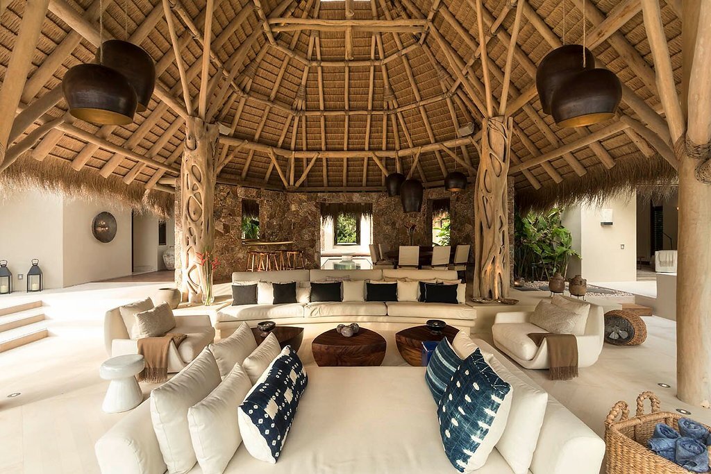 Grand palapa living pavilion with carved wood columns, a soaring thatched ceiling, a curved cream sectional with indigo ikat pillows, two drum pendant lights, and stone accent walls, Casa Escondida, Ranchos Estates, Punta Mita.
