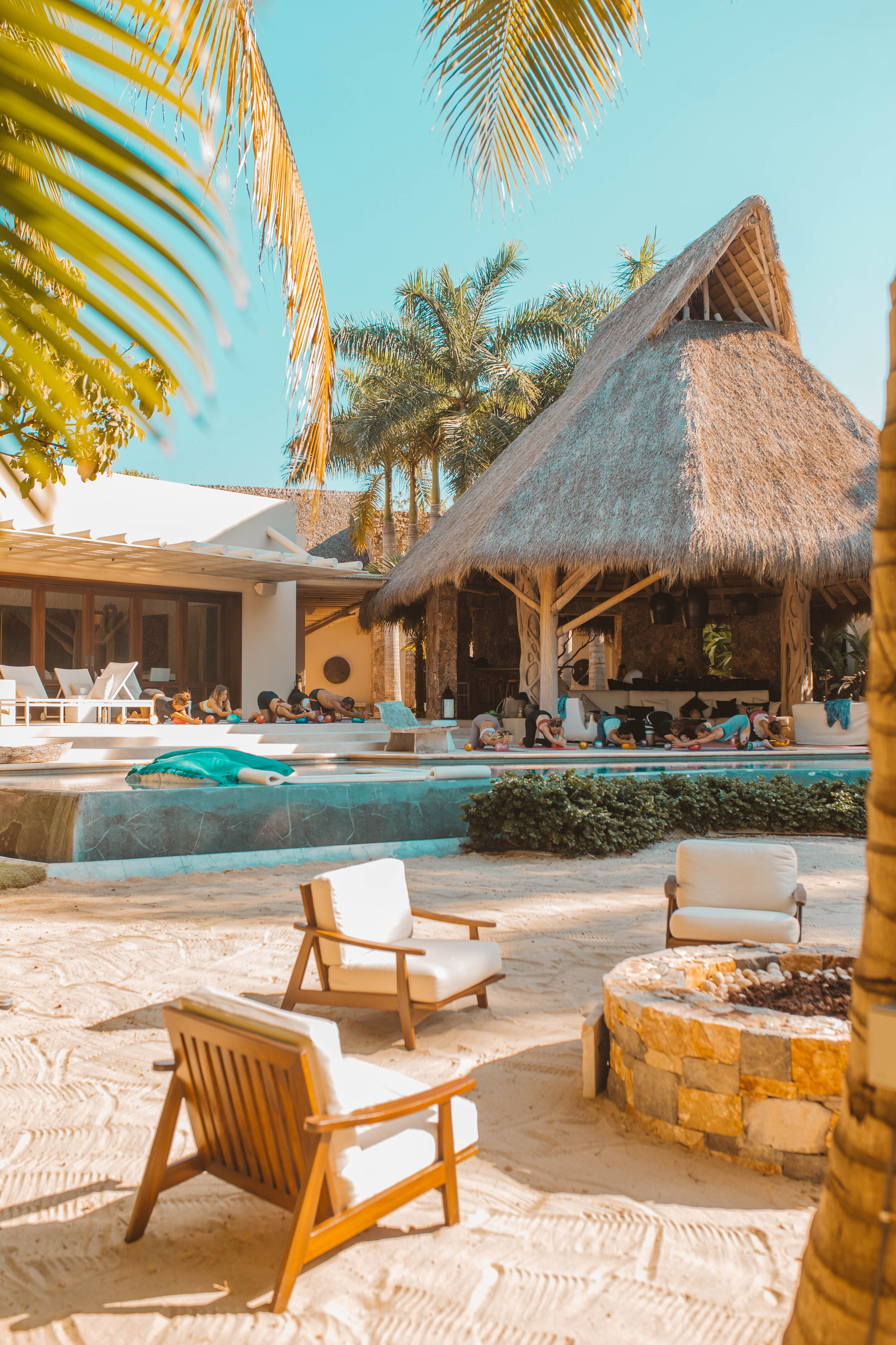 View from the sandy beach side across teak armchairs around a stone fire pit toward the pool, palapa bar beyond, and the villa's cream pavilion wings framed by palm trees, Casa Escondida, Ranchos Estates, Punta Mita.