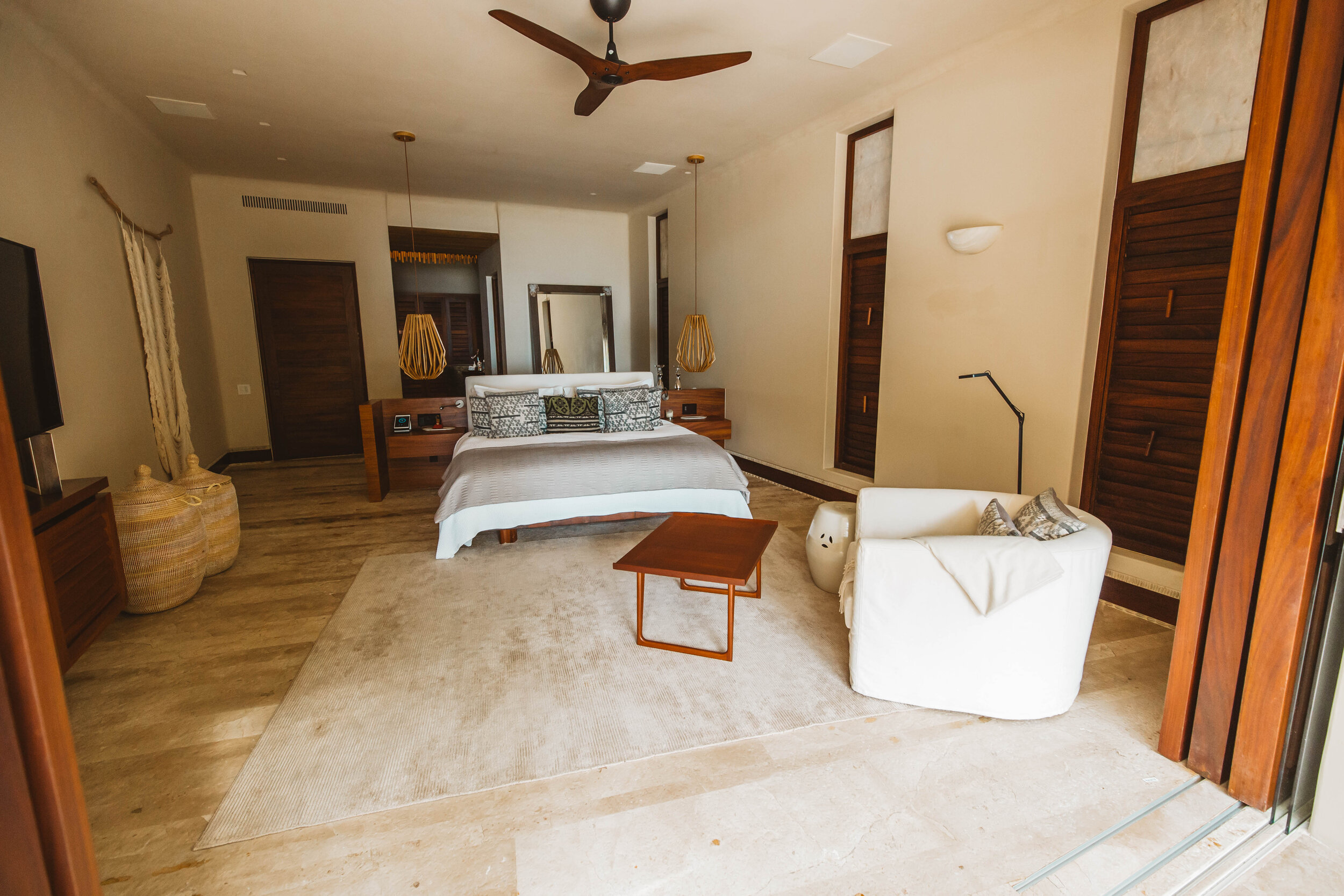 Spacious primary suite seen from the open terrace doorway, king platform bed with woven pendant lights above, louvered shutters on three walls, ceiling fan, and large woven baskets flanking a credenza with a flat screen, Casa Escondida, Ranchos Estates, Punta Mita.