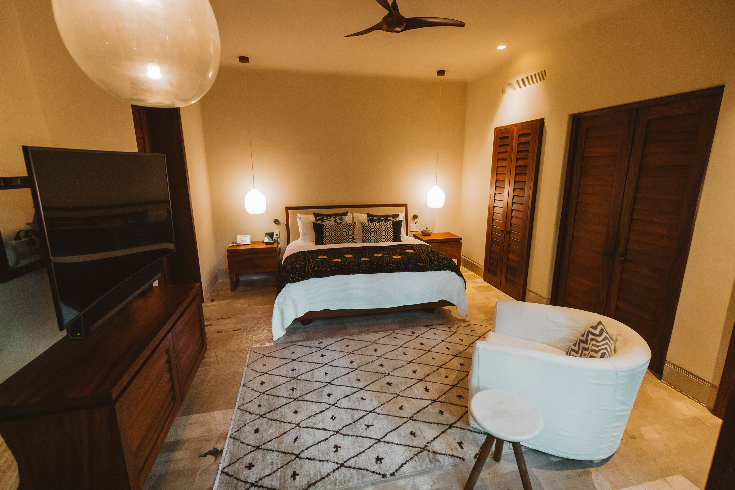 Guest suite at night with a king bed dressed in a dark batik throw, louvered closet doors, globe pendant lights, a diamond-pattern Berber rug, and a large flat-screen television on a dark wood console, Casa Escondida, Ranchos Estates, Punta Mita.