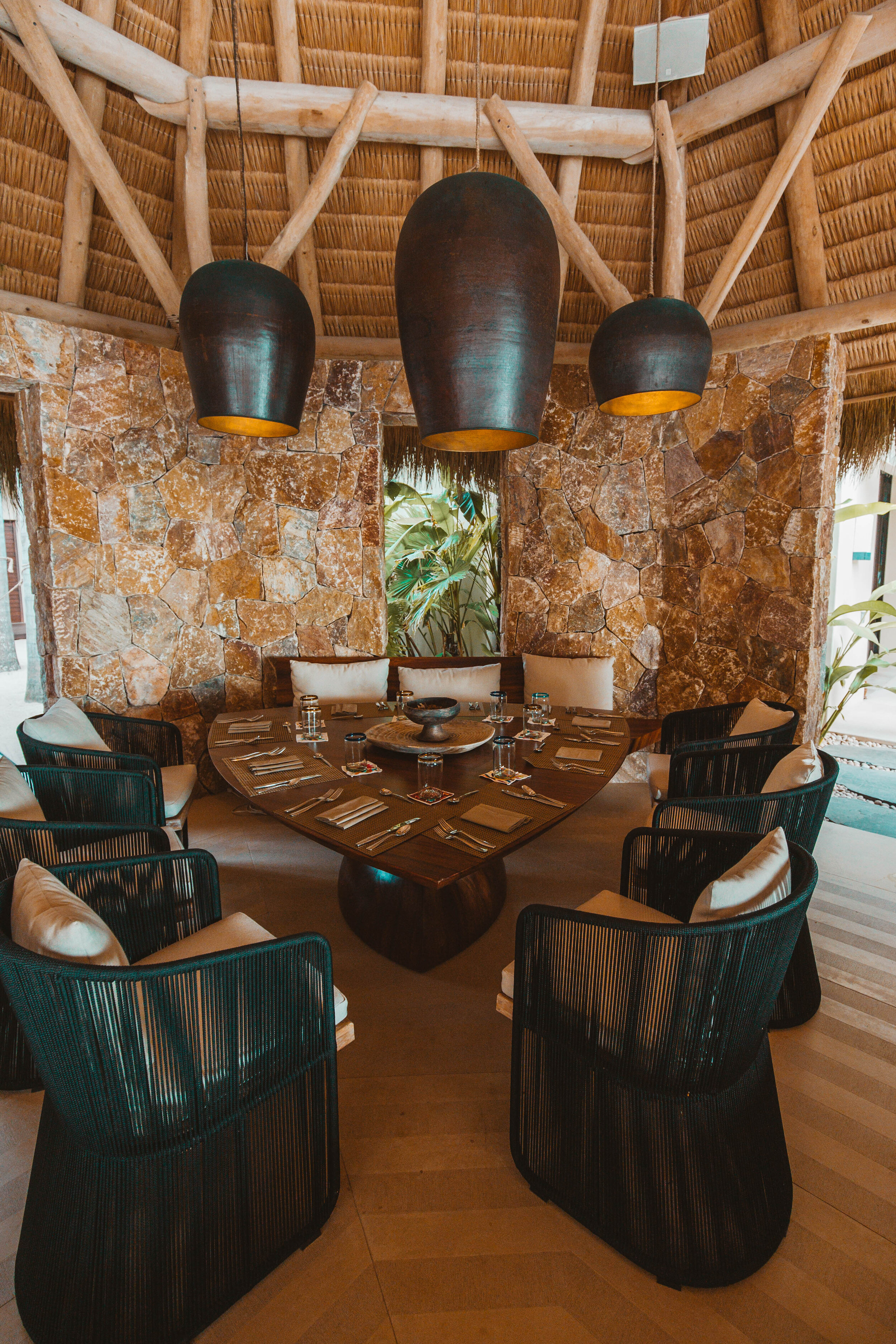 Palapa dining room with a long dark wood table set for ten, dark rope-woven chairs, three oversized drum pendants, and a rough-cut stone feature wall, Casa Escondida, Ranchos Estates, Punta Mita.