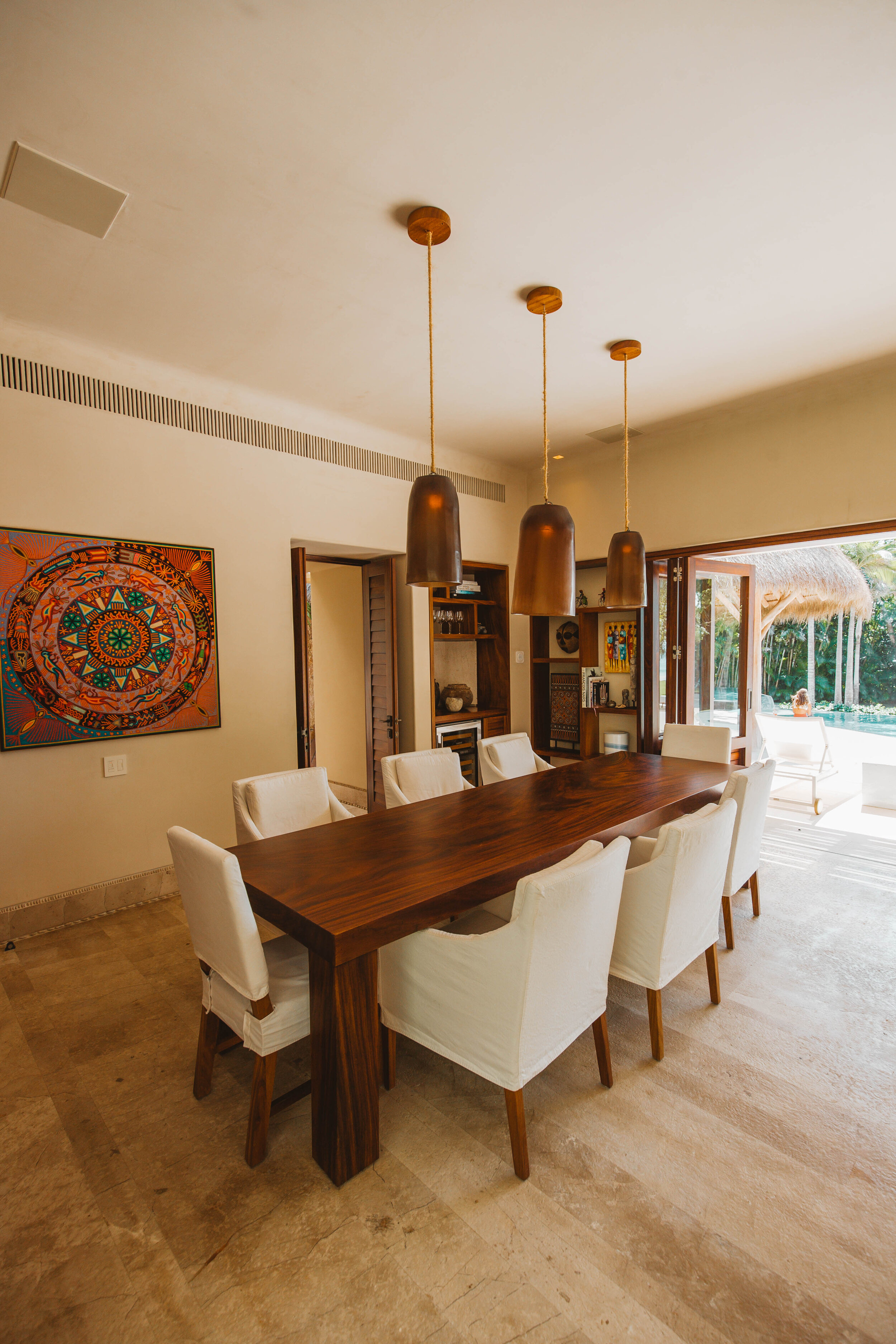 Interior dining room with a long walnut slab table, eight white slipcover chairs, three bronze drop pendants, a vibrant Huichol mandala canvas on the wall, and sliding doors open to the pool terrace, Casa Escondida, Ranchos Estates, Punta Mita.