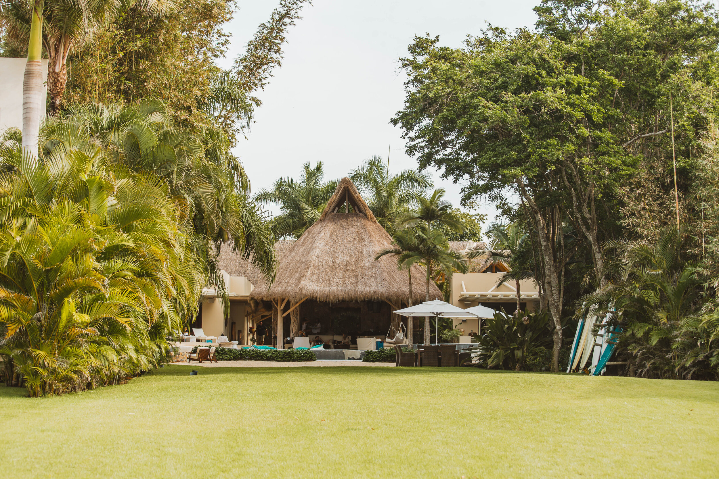 Arrival lawn view of the villa from the garden, palapa lounge at center, cream stucco wings on each side, lounge chairs and surfboards visible, all framed by tall palms and tropical trees, Casa Escondida, Ranchos Estates, Punta Mita.