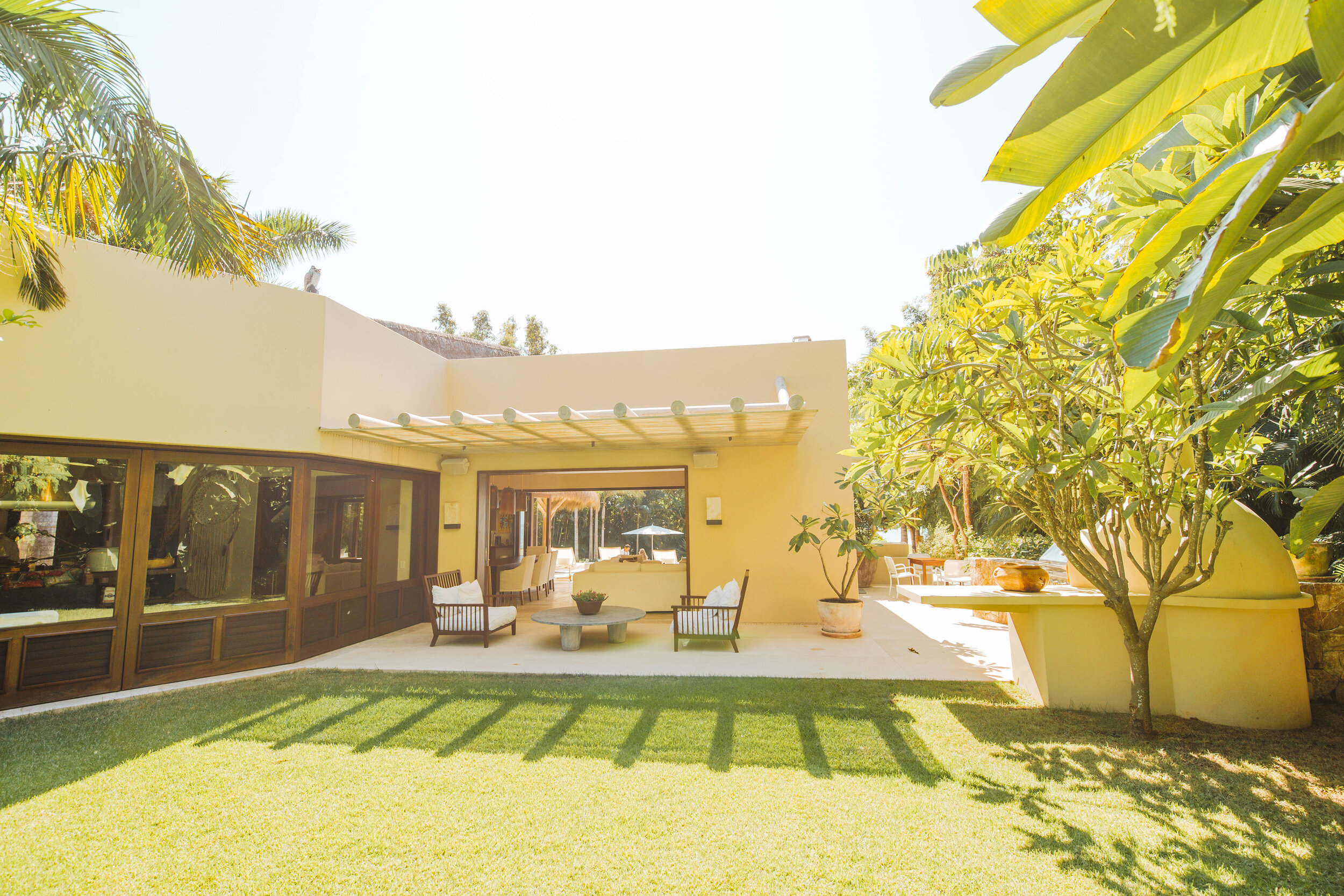 Garden-side terrace with cream stucco walls, wood-framed sliding doors, a white pergola, patio furniture on a stone terrace, and a plumeria tree beside a terracotta planter, Casa Escondida, Ranchos Estates, Punta Mita.