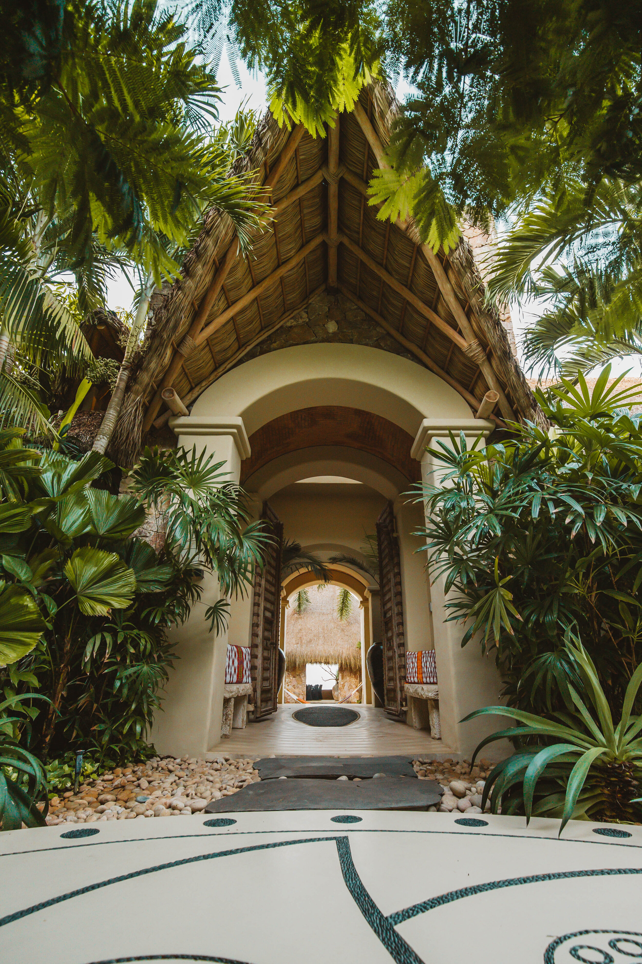 Arched entrance corridor of repeating white arches with a palapa thatch roof above, a mosaic stone path underfoot, Otomi textile panels hanging in the doorways, and dense tropical palms on both sides, Casa Escondida, Ranchos Estates, Punta Mita.