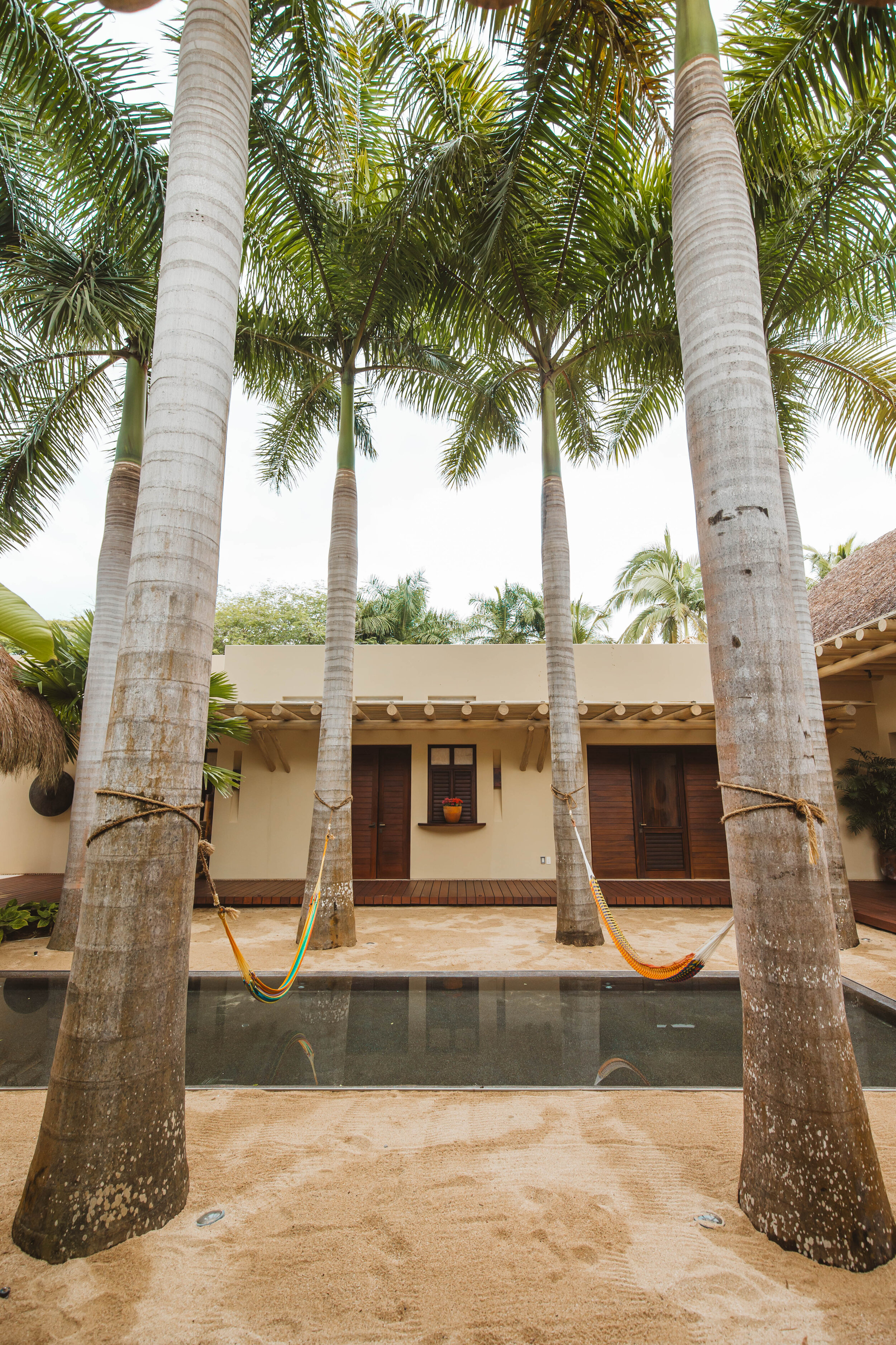 Courtyard reflecting pool seen from the far end, four royal palms with colorful hammocks strung between them over the sand, cream stucco guest wing and louvered shutters in the background, Casa Escondida, Ranchos Estates, Punta Mita.