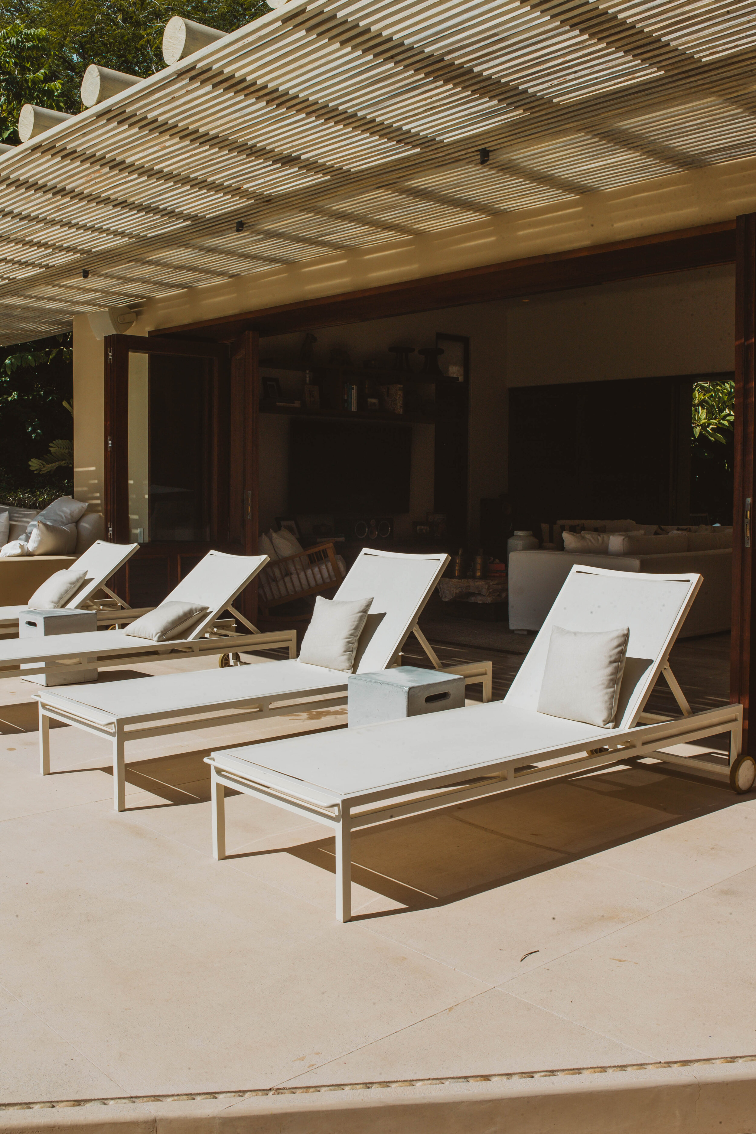 Three white pool loungers lined up under a slatted palapa terrace, the open living pavilion visible in shade behind them, Casa Escondida, Ranchos Estates, Punta Mita.