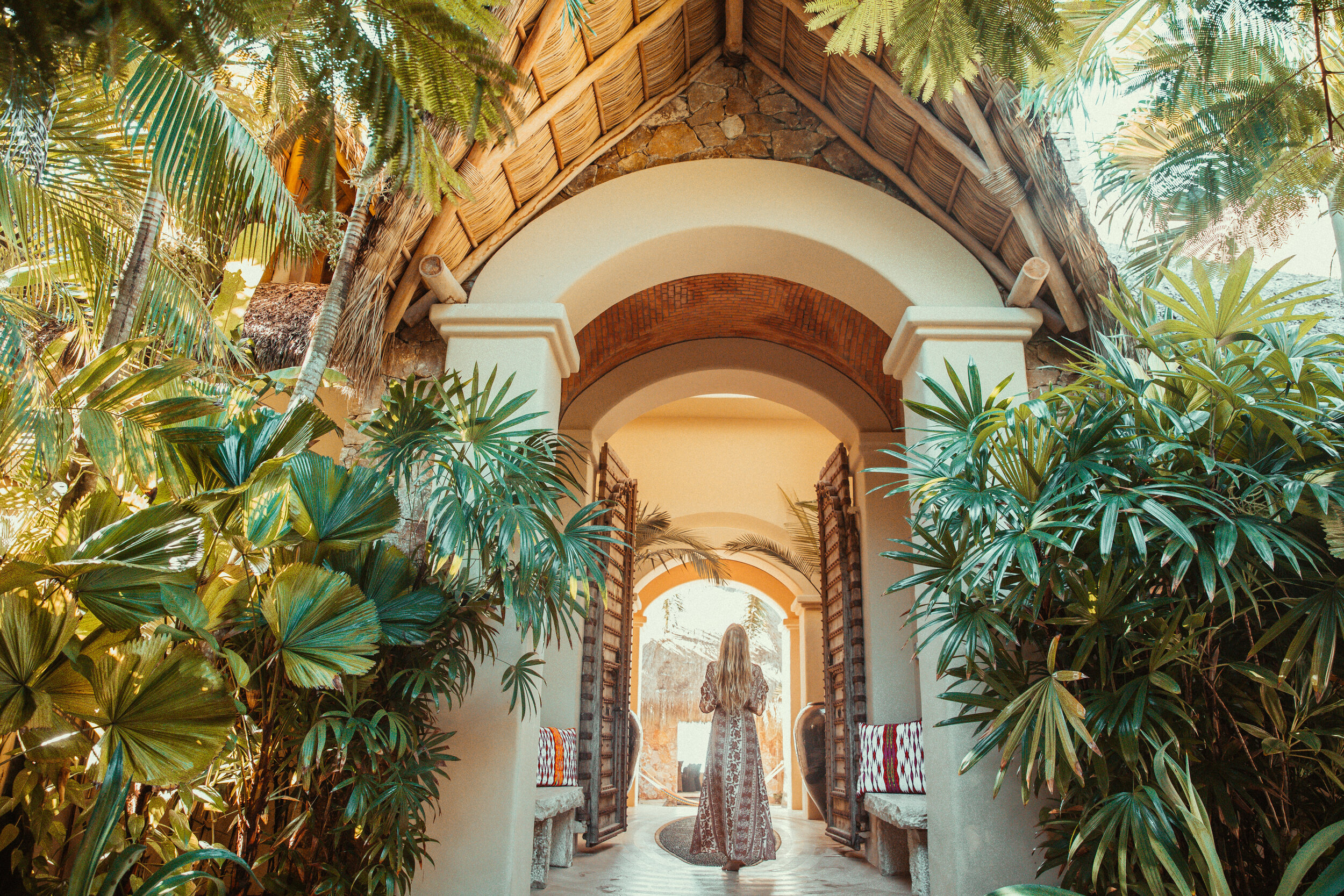 A guest walks through the arched stone entry corridor lined with carved wood doors and fan palms, the inner courtyard glowing at the far end, Casa Escondida, Ranchos Estates, Punta Mita.